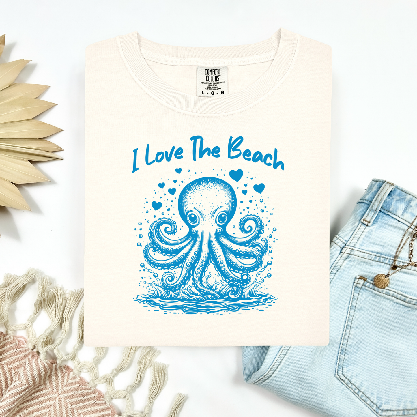The Comfort Colors 1717 "I Love The Beach" T-shirt features a charming one-color graphic of an octopus surrounded by hearts, bringing a touch of whimsy and coastal charm. Displayed in a flat lay mockup, the tee is styled with a pair of blue jeans, a cozy blanket, and a wicker sun-shaped accessory—perfect for showcasing a relaxed, beachy vibe.