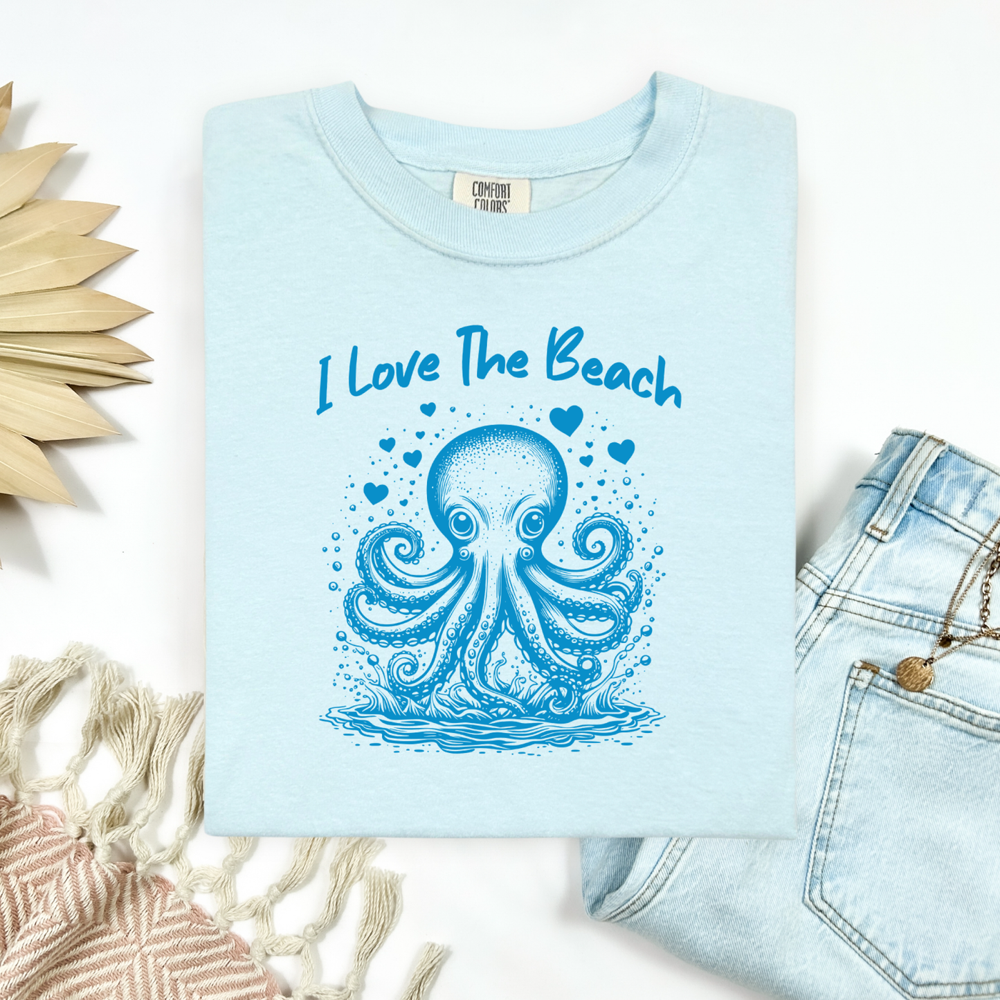 The Comfort Colors 1717 "I Love The Beach" T-shirt features a charming one-color graphic of an octopus surrounded by hearts, bringing a touch of whimsy and coastal charm. Displayed in a flat lay mockup, the tee is styled with a pair of blue jeans, a cozy blanket, and a wicker sun-shaped accessory—perfect for showcasing a relaxed, beachy vibe.