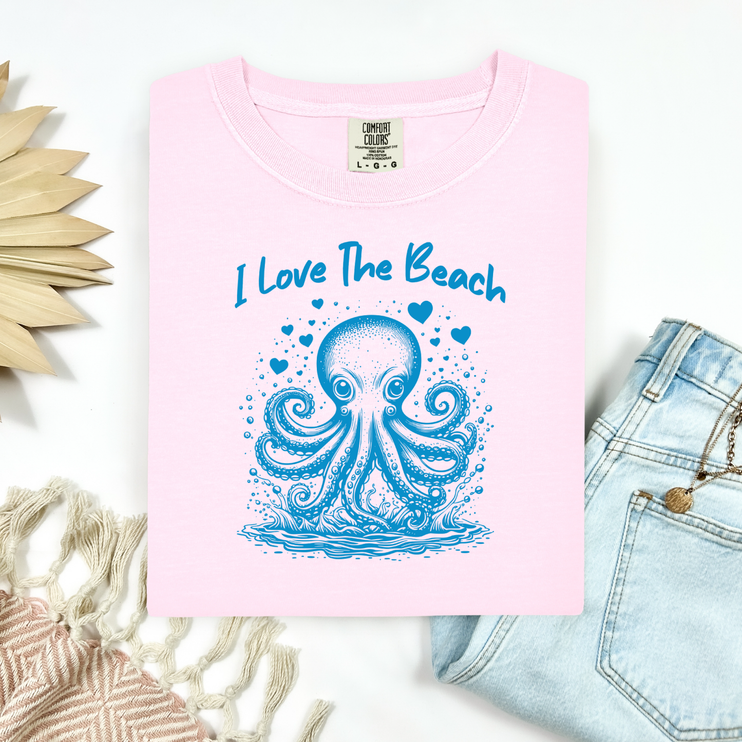The Comfort Colors 1717 "I Love The Beach" T-shirt features a charming one-color graphic of an octopus surrounded by hearts, bringing a touch of whimsy and coastal charm. Displayed in a flat lay mockup, the tee is styled with a pair of blue jeans, a cozy blanket, and a wicker sun-shaped accessory—perfect for showcasing a relaxed, beachy vibe.