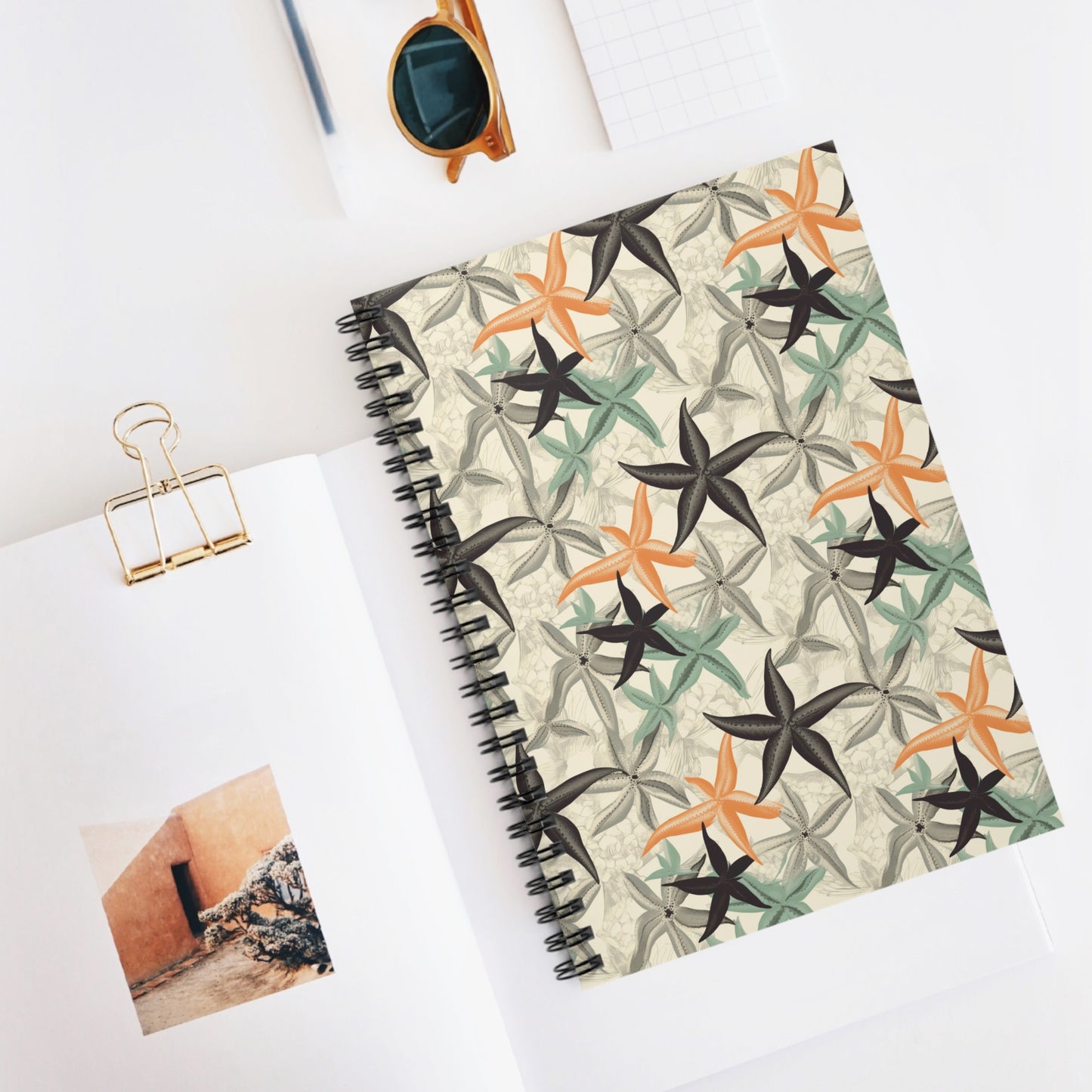 The Tropical Starfish Spiral Notebook, featuring a starfish pattern cover, lies open on a white surface with nature-inspired charm. Nearby, a small notepad rests next to sunglasses and an open book showcasing an orange building adorned with a gold binder clip.