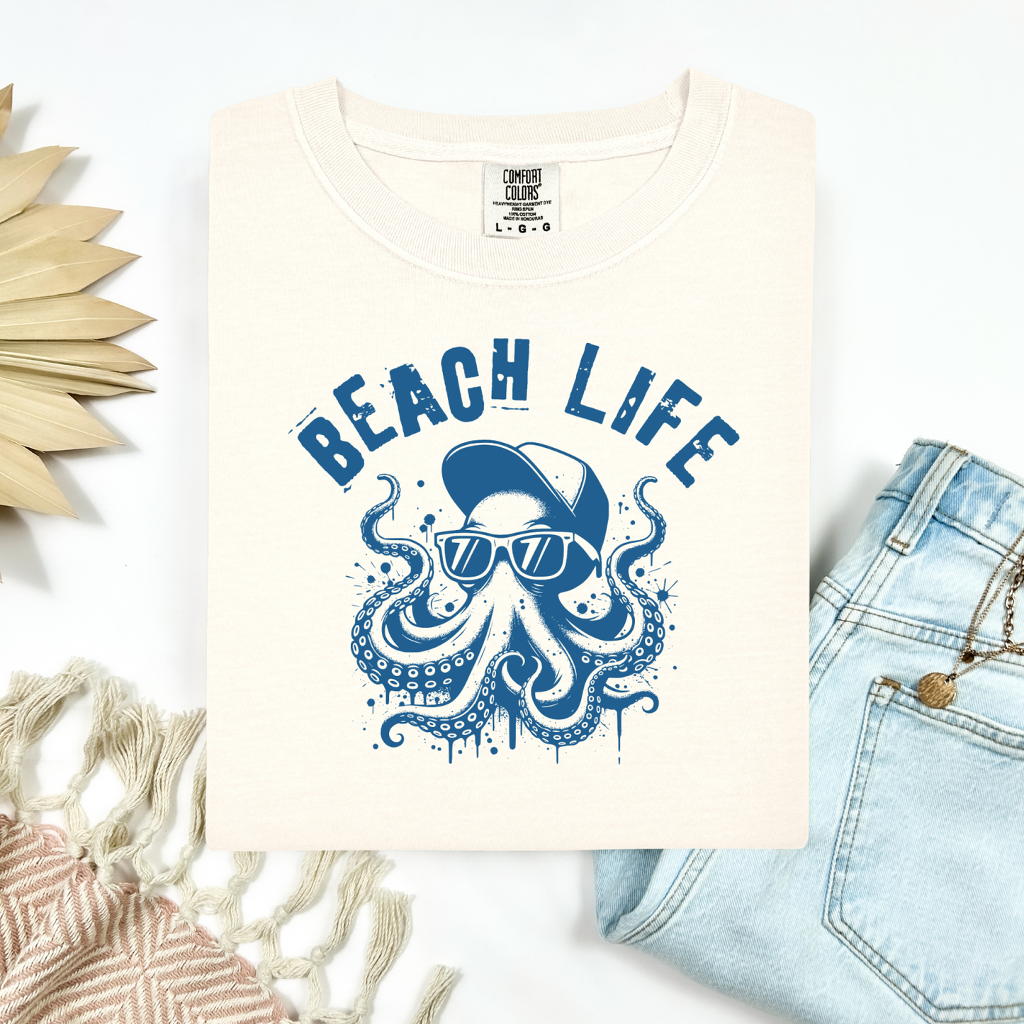 Comfort Colors 1717 "Beach Life" t shirt with a octopus with a baseball cap and sunglasses in a one color graphic. Picture on a flay lay mockup with pair of blue jeans and blanket and a wicker sun shaped accessory.