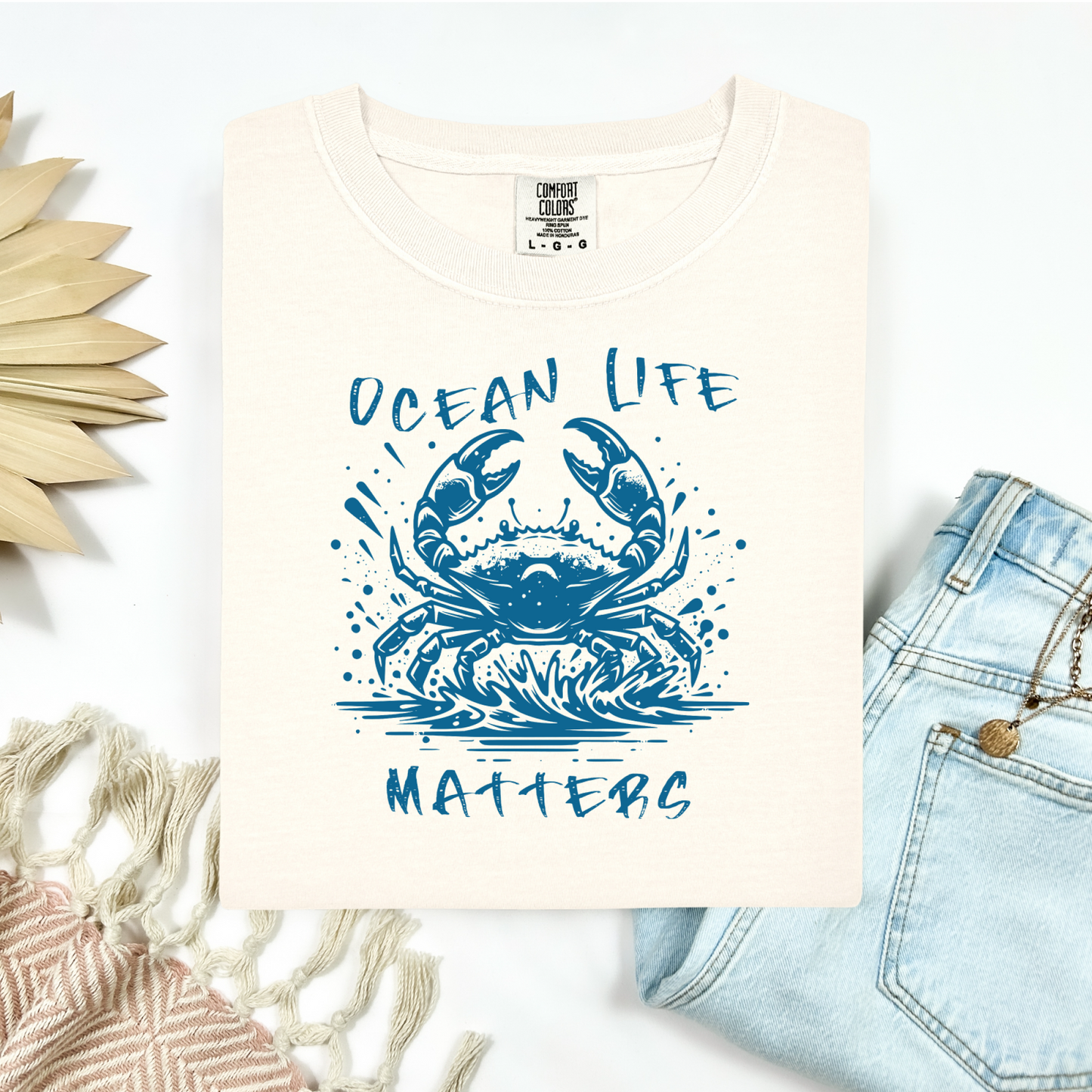 Comfort Colors 1717 "Ocean Life Matters" t shirt with a crab splashing in water in a one color graphic. Picture on a flay lay mockup with pair of blue jeans and blanket and a wicker sun shaped accessory.