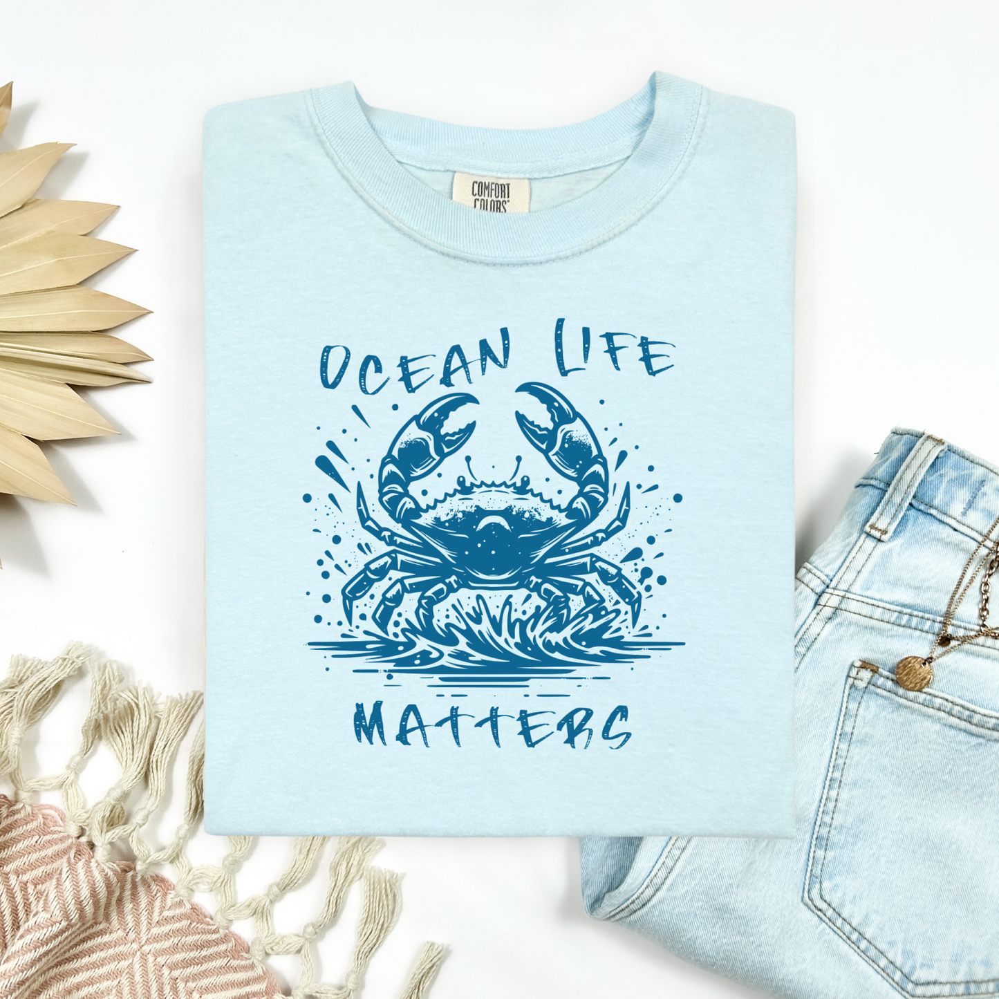 Comfort Colors 1717 "Ocean Life Matters" t shirt with a crab splashing in water in a one color graphic. Picture on a flay lay mockup with pair of blue jeans and blanket and a wicker sun shaped accessory.