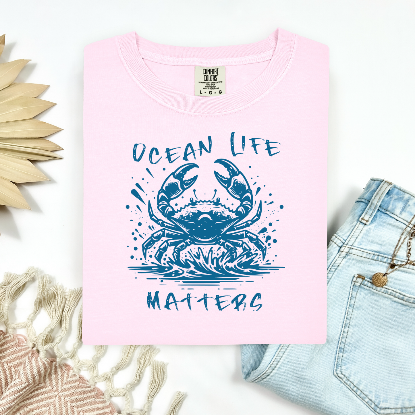 Comfort Colors 1717 "Ocean Life Matters" t shirt with a crab splashing in water in a one color graphic. Picture on a flay lay mockup with pair of blue jeans and blanket and a wicker sun shaped accessory.