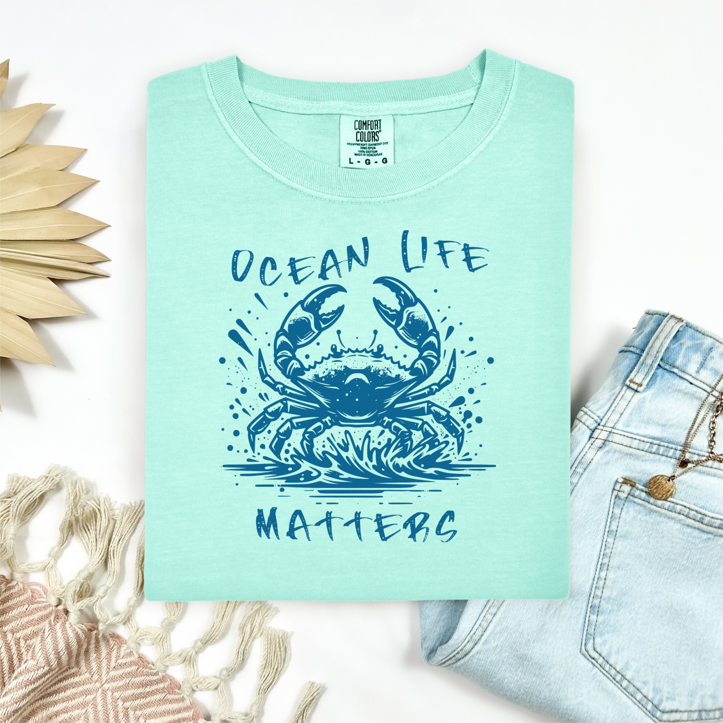 Comfort Colors 1717 "Ocean Life Matters" t shirt with a crab splashing in water in a one color graphic. Picture on a flay lay mockup with pair of blue jeans and blanket and a wicker sun shaped accessory.