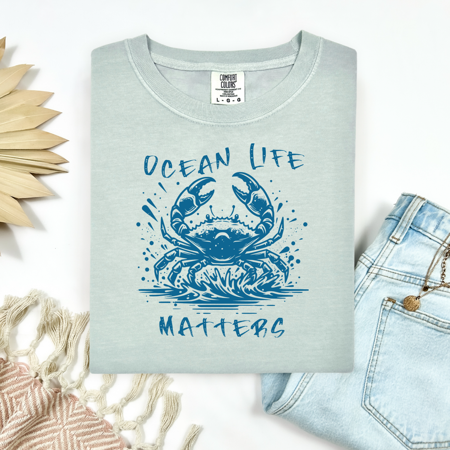 Comfort Colors 1717 "Ocean Life Matters" t shirt with a crab splashing in water in a one color graphic. Picture on a flay lay mockup with pair of blue jeans and blanket and a wicker sun shaped accessory.
