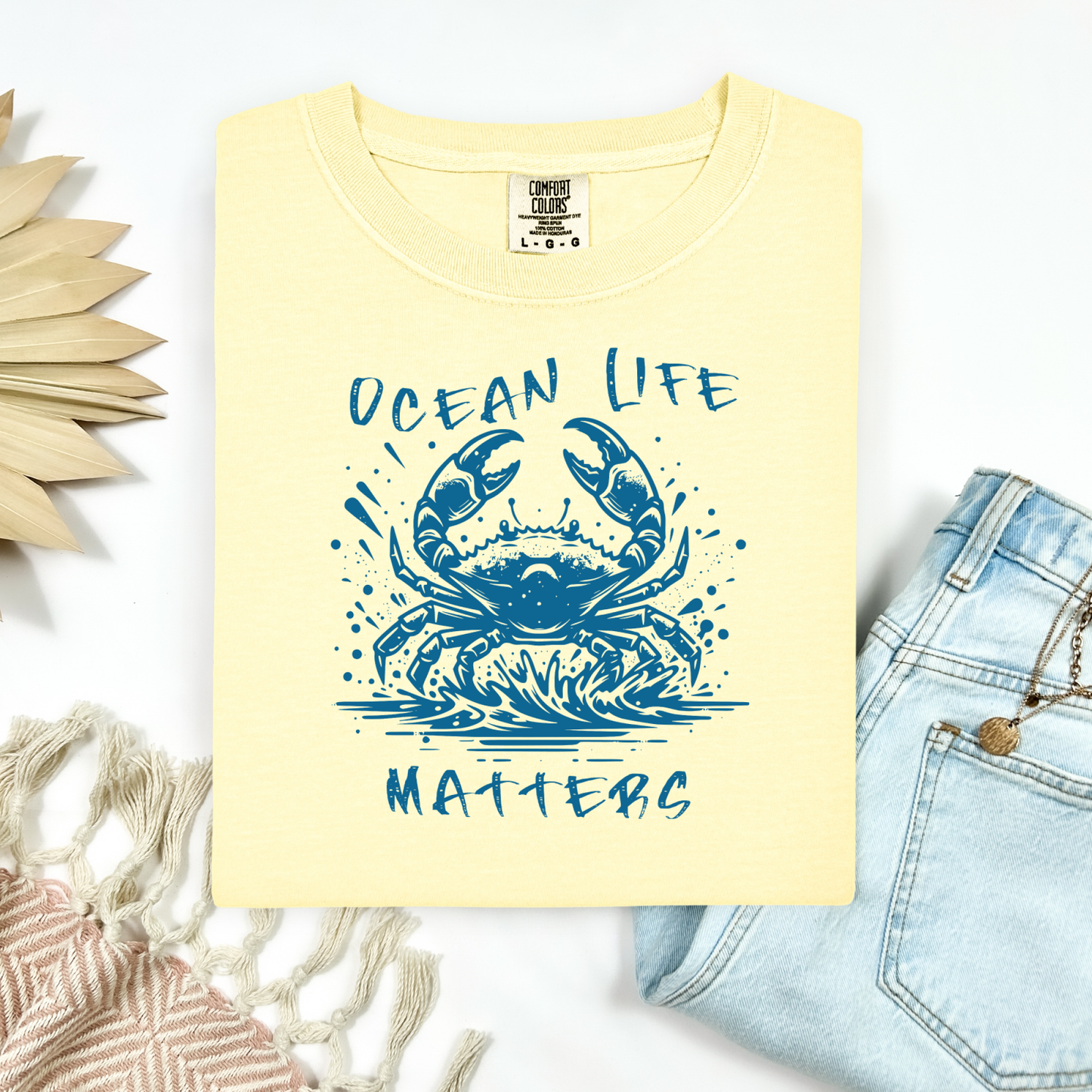 Comfort Colors 1717 "Ocean Life Matters" t shirt with a crab splashing in water in a one color graphic. Picture on a flay lay mockup with pair of blue jeans and blanket and a wicker sun shaped accessory.