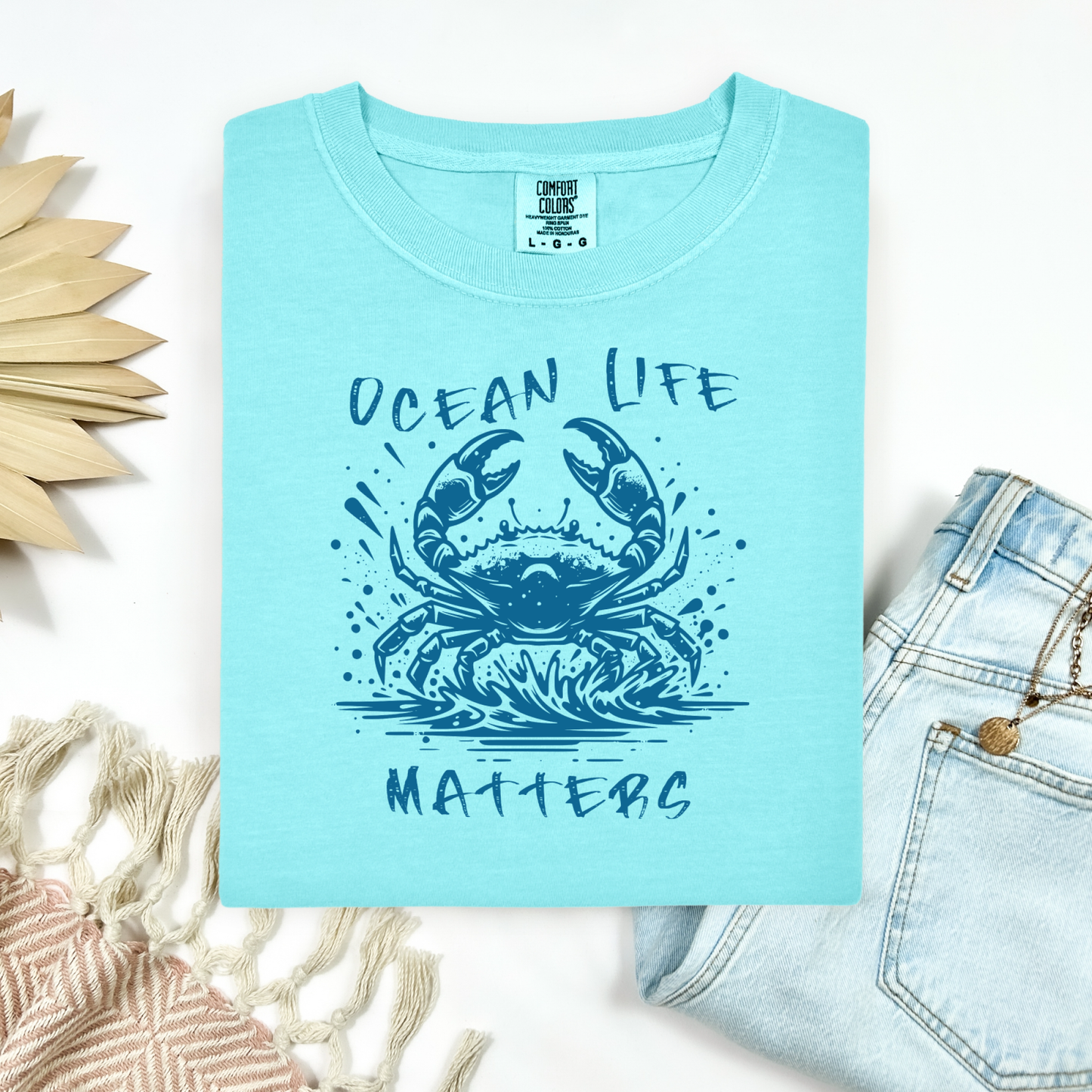 Comfort Colors 1717 "Ocean Life Matters" t shirt with a crab splashing in water in a one color graphic. Picture on a flay lay mockup with pair of blue jeans and blanket and a wicker sun shaped accessory.