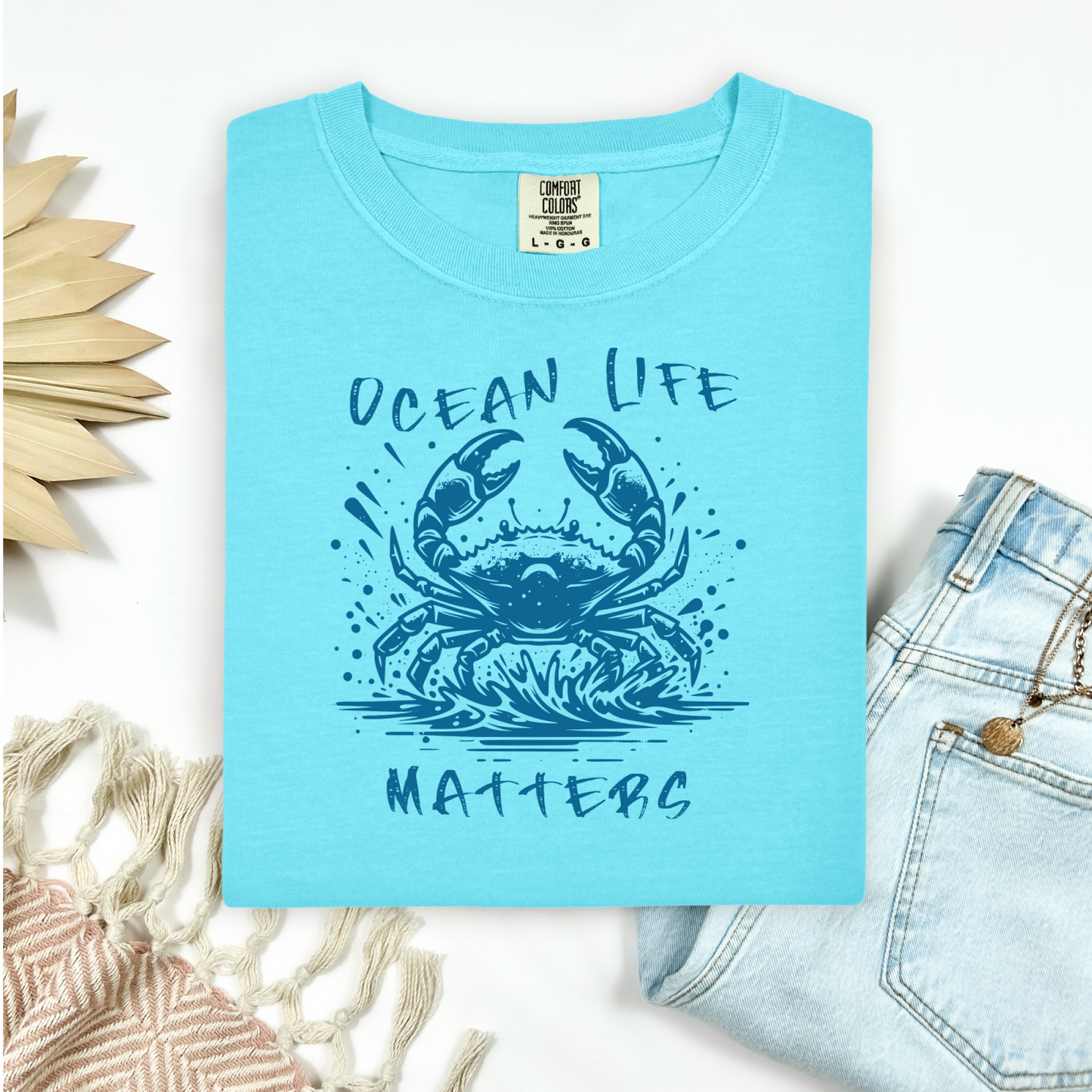 Comfort Colors 1717 "Ocean Life Matters" t shirt with a crab splashing in water in a one color graphic. Picture on a flay lay mockup with pair of blue jeans and blanket and a wicker sun shaped accessory.