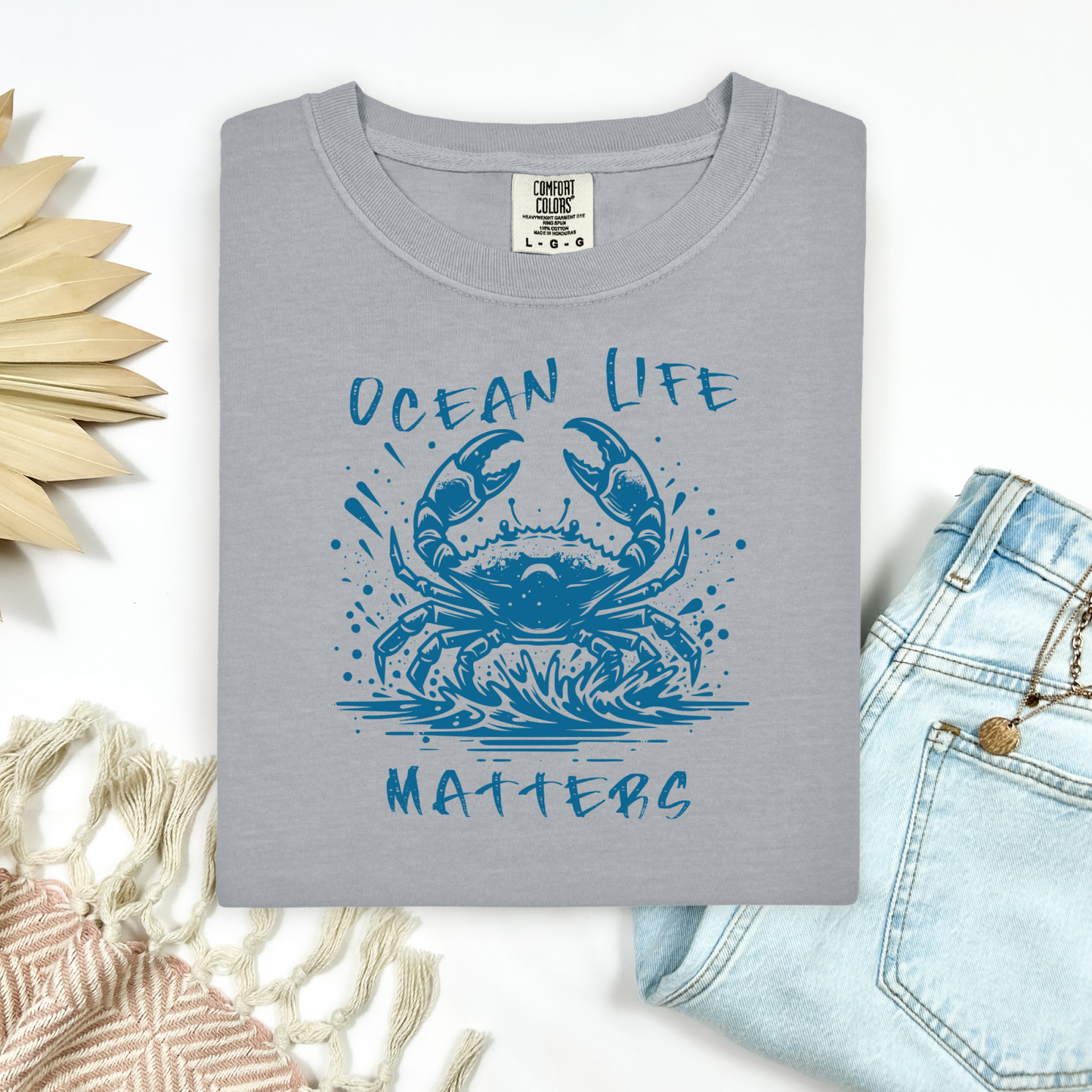 Comfort Colors 1717 "Ocean Life Matters" t shirt with a crab splashing in water in a one color graphic. Picture on a flay lay mockup with pair of blue jeans and blanket and a wicker sun shaped accessory.