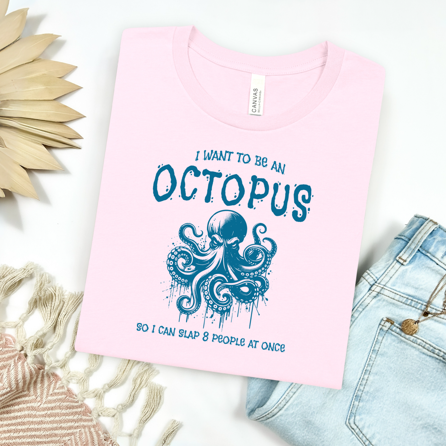 Bella Canvas 3001 "I Want To Be An Octopus So I Can Slap 8 People At Once" t shirt with an angry octopus one color graphic. Picture on a flay lay mockup with pair of blue jeans and blanket and a wicker sun shaped accessory.