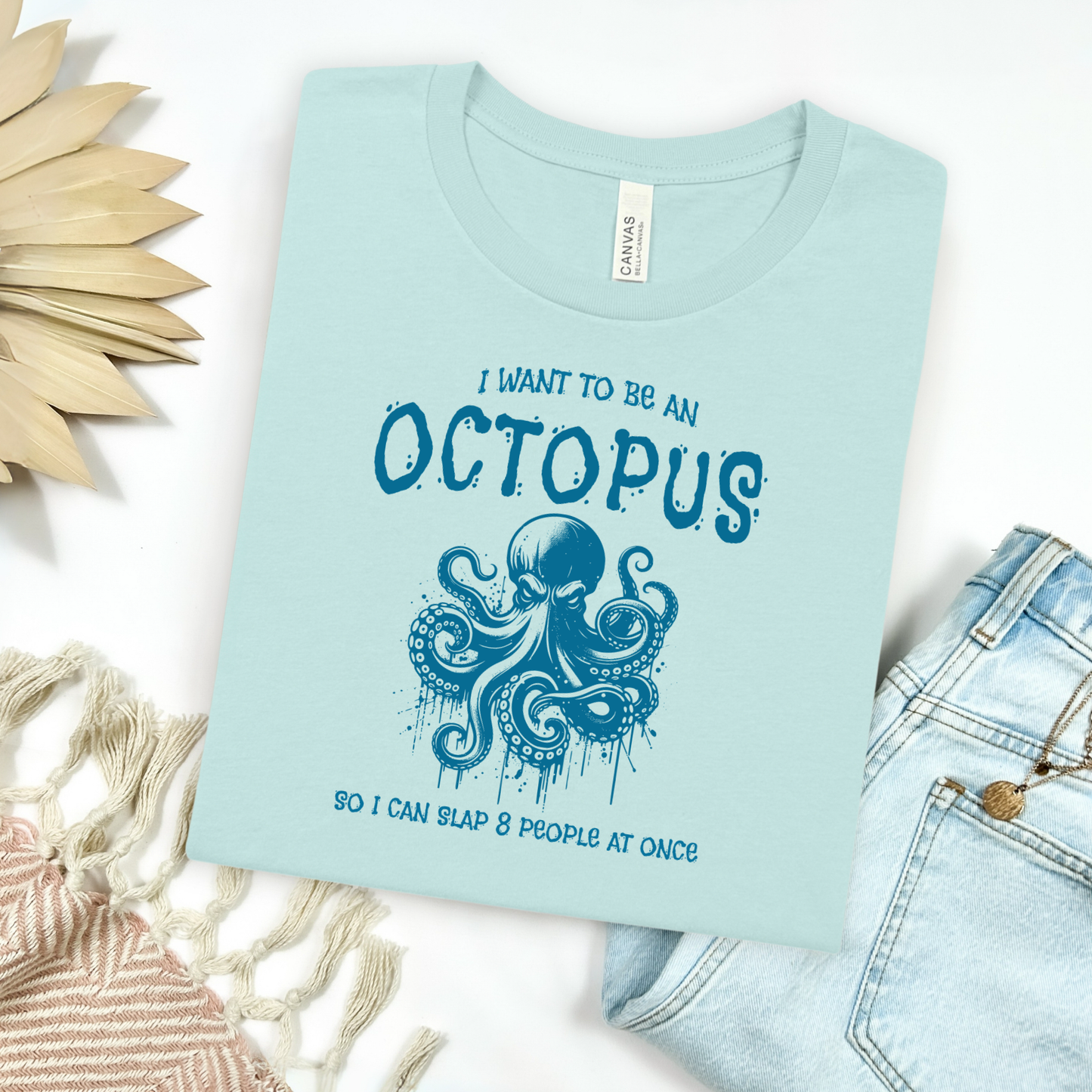Bella Canvas 3001 "I Want To Be An Octopus So I Can Slap 8 People At Once" t shirt with an angry octopus one color graphic. Picture on a flay lay mockup with pair of blue jeans and blanket and a wicker sun shaped accessory.