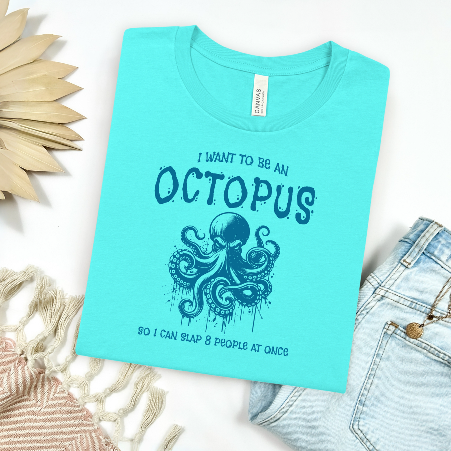 Bella Canvas 3001 "I Want To Be An Octopus So I Can Slap 8 People At Once" t shirt with an angry octopus one color graphic. Picture on a flay lay mockup with pair of blue jeans and blanket and a wicker sun shaped accessory.