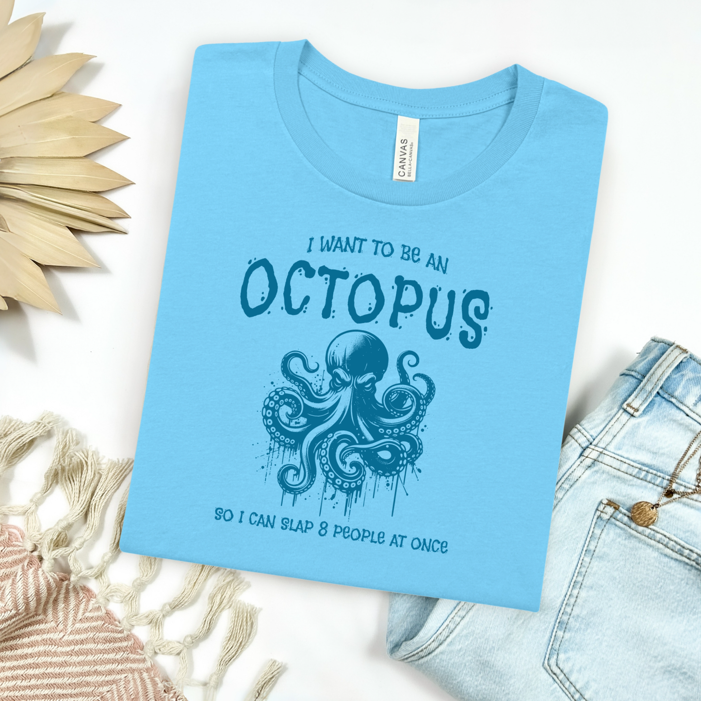 Bella Canvas 3001 "I Want To Be An Octopus So I Can Slap 8 People At Once" t shirt with an angry octopus one color graphic. Picture on a flay lay mockup with pair of blue jeans and blanket and a wicker sun shaped accessory.
