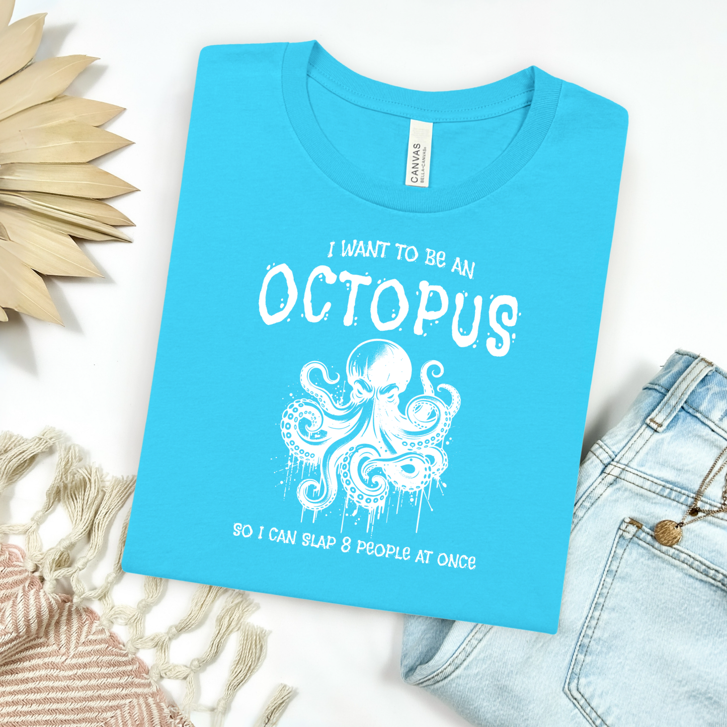 Bella Canvas 3001 "I Want To Be An Octopus So I Can Slap 8 People At Once" t shirt with an angry octopus one color graphic. Picture on a flay lay mockup with pair of blue jeans and blanket and a wicker sun shaped accessory.