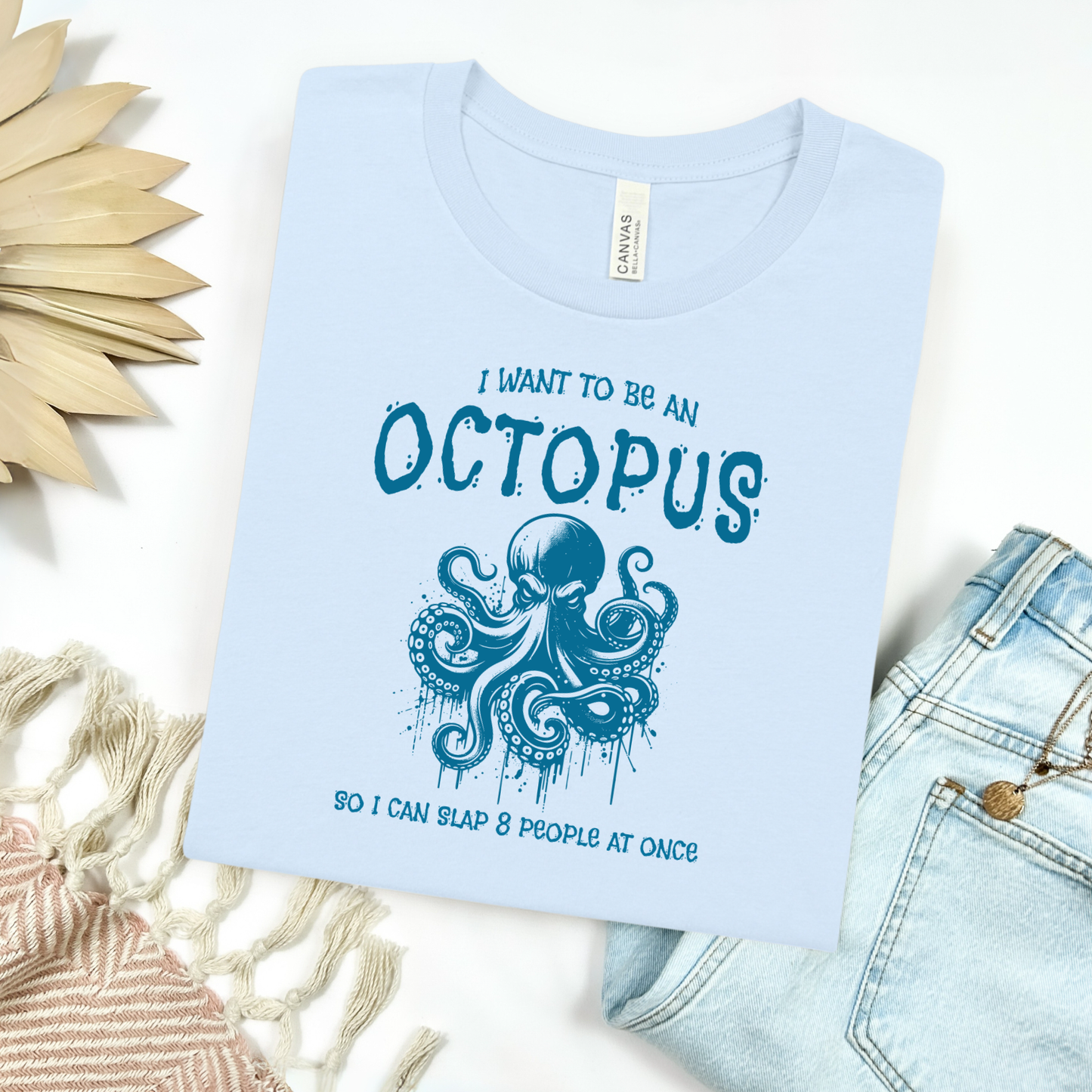 Bella Canvas 3001 "I Want To Be An Octopus So I Can Slap 8 People At Once" t shirt with an angry octopus one color graphic. Picture on a flay lay mockup with pair of blue jeans and blanket and a wicker sun shaped accessory.