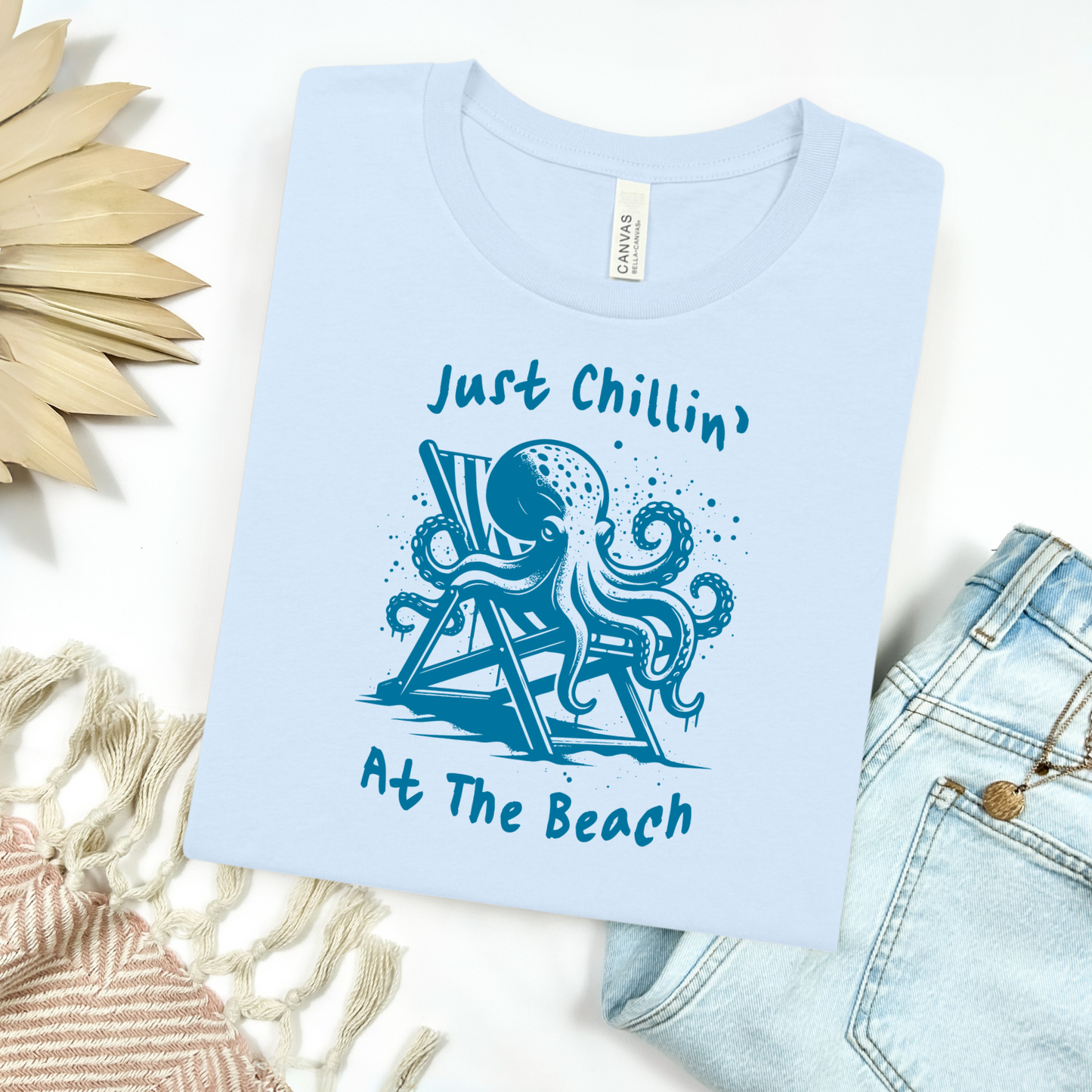 Bella Canvas 3001 "Just Chillin' At The Beach" t shirt with an octopus lounging in a beach chair in a one color graphic. Picture on a flay lay mockup with pair of blue jeans and blanket and a wicker sun shaped accessory.