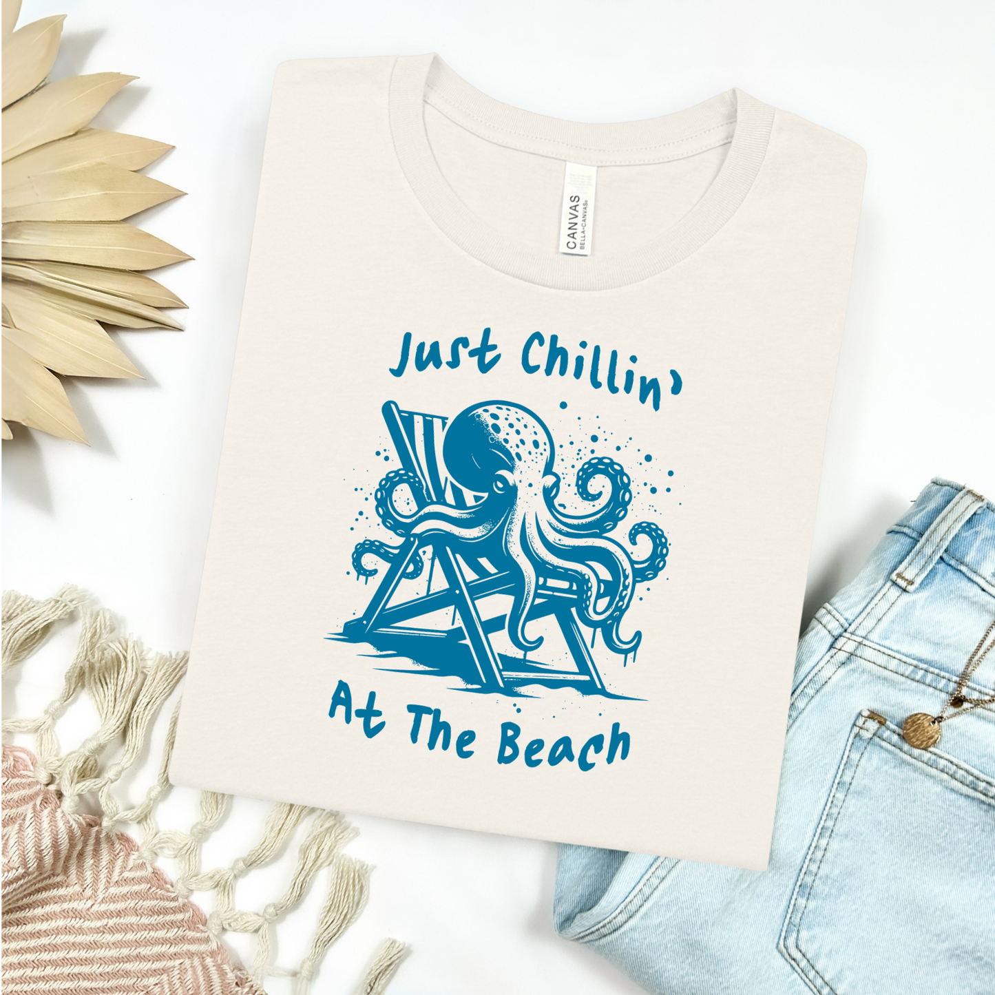 Bella Canvas 3001 "Just Chillin' At The Beach" t shirt with an octopus lounging in a beach chair in a one color graphic. Picture on a flay lay mockup with pair of blue jeans and blanket and a wicker sun shaped accessory.