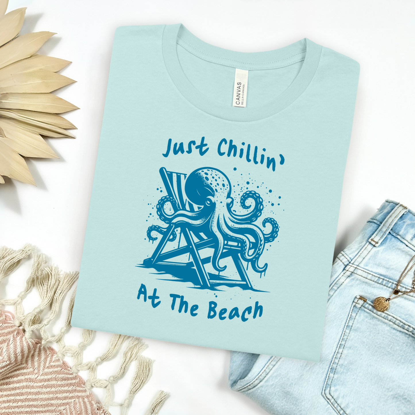 Bella Canvas 3001 "Just Chillin' At The Beach" t shirt with an octopus lounging in a beach chair in a one color graphic. Picture on a flay lay mockup with pair of blue jeans and blanket and a wicker sun shaped accessory.