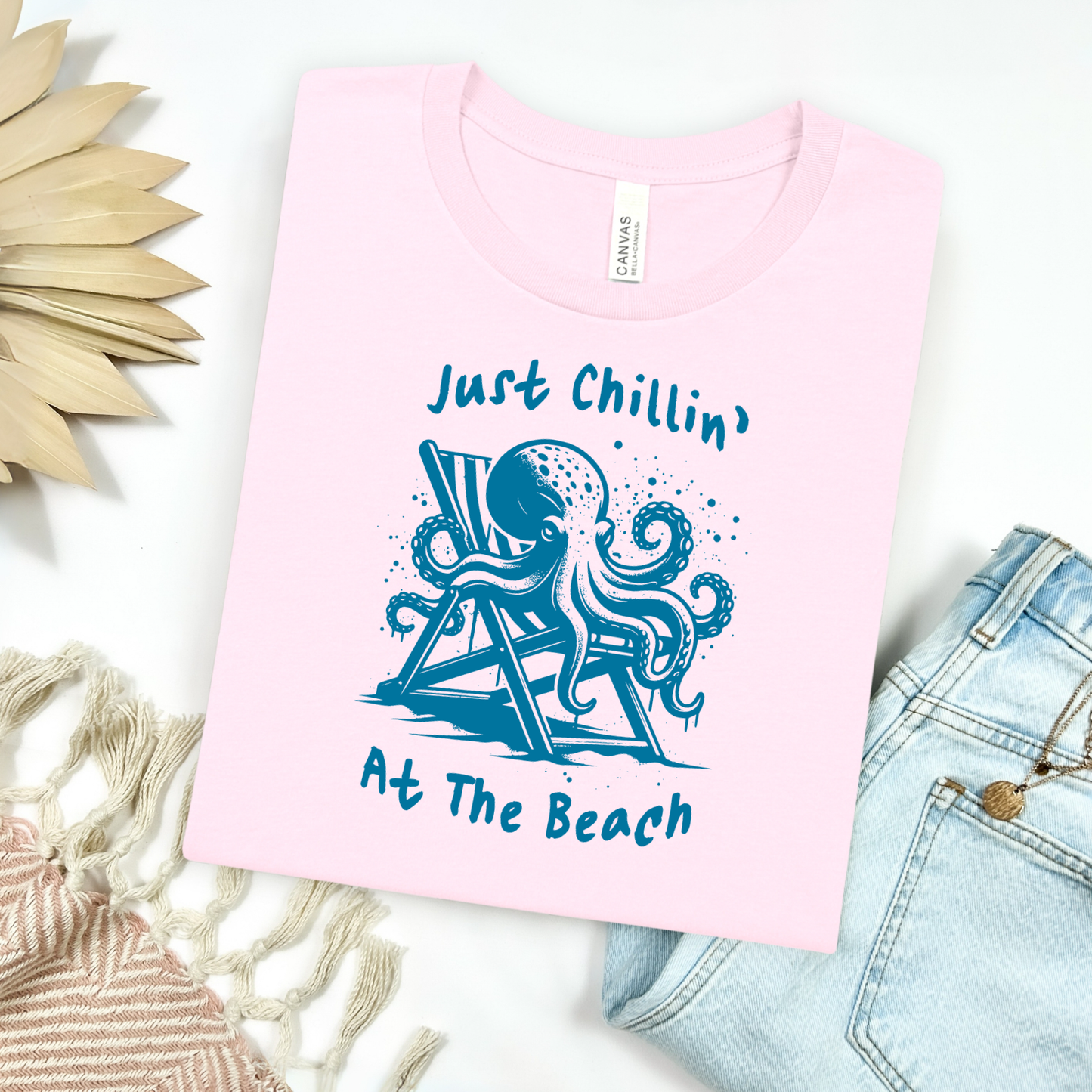 Bella Canvas 3001 "Just Chillin' At The Beach" t shirt with an octopus lounging in a beach chair in a one color graphic. Picture on a flay lay mockup with pair of blue jeans and blanket and a wicker sun shaped accessory.