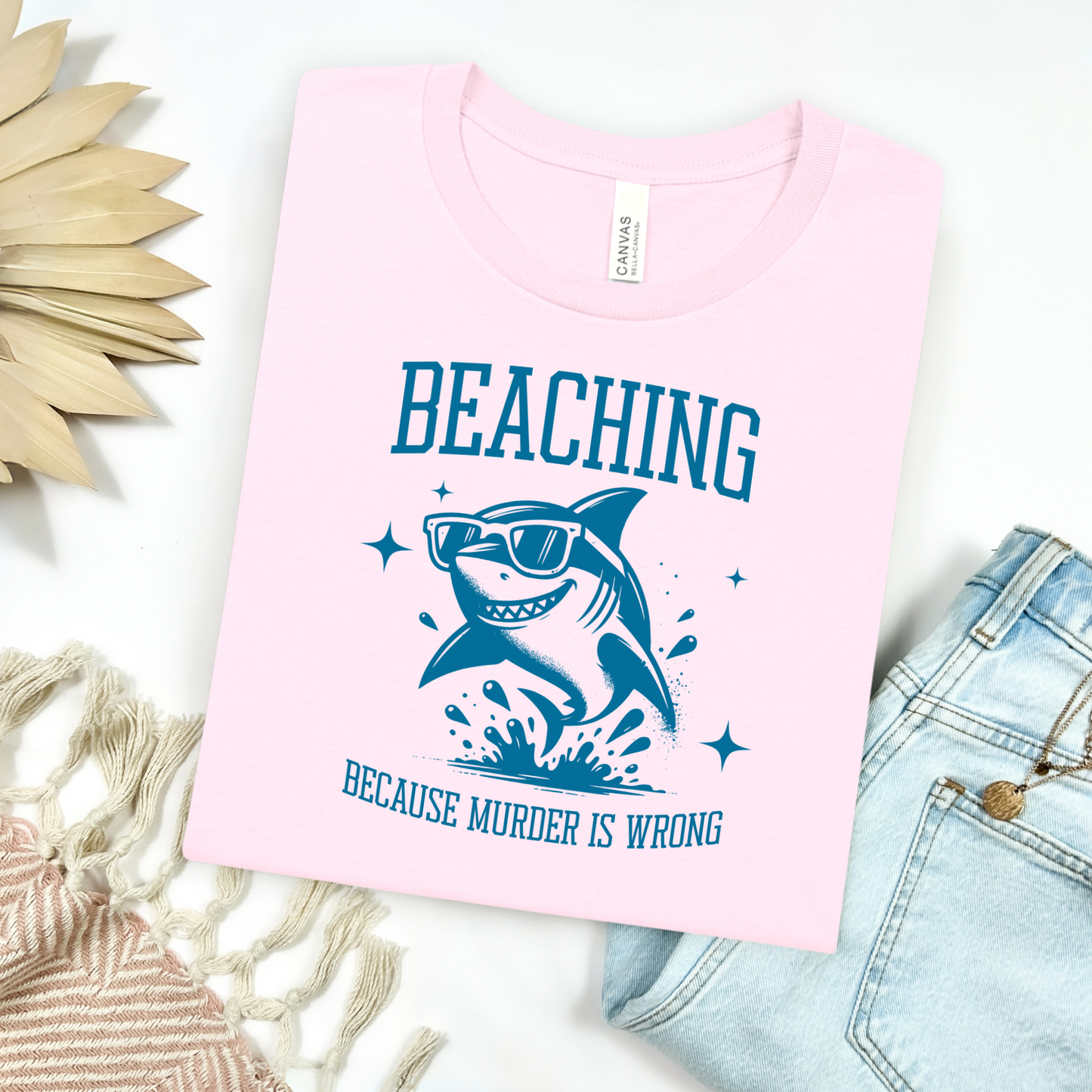 Bella Canvas 3001 Beaching Because Murder Is Wrong T Shirt with Funny Shark with Sunglasses splashing in water in one color graphic. Picture on a flay lay mockup with pair of blue jeans and blanket and a wicker sun shaped accessory.