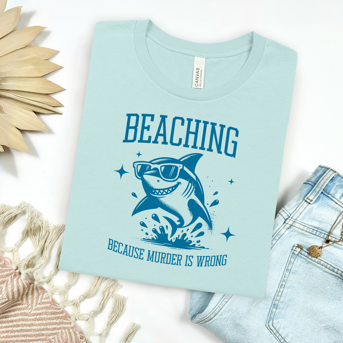 Bella Canvas 3001 Beaching Because Murder Is Wrong T Shirt with Funny Shark with Sunglasses splashing in water in one color graphic. Picture on a flay lay mockup with pair of blue jeans and blanket and a wicker sun shaped accessory.