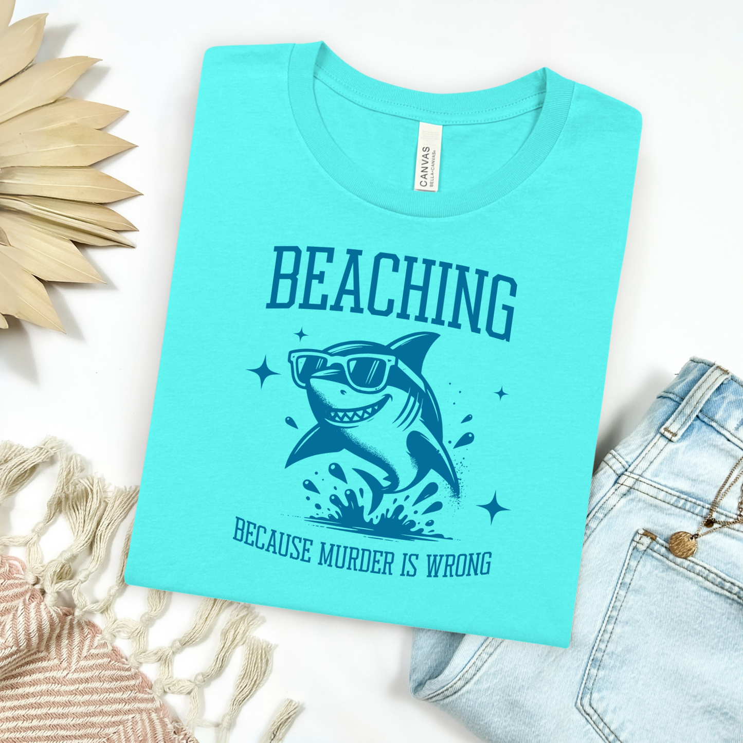 Bella Canvas 3001 Beaching Because Murder Is Wrong T Shirt with Funny Shark with Sunglasses splashing in water in one color graphic. Picture on a flay lay mockup with pair of blue jeans and blanket and a wicker sun shaped accessory.