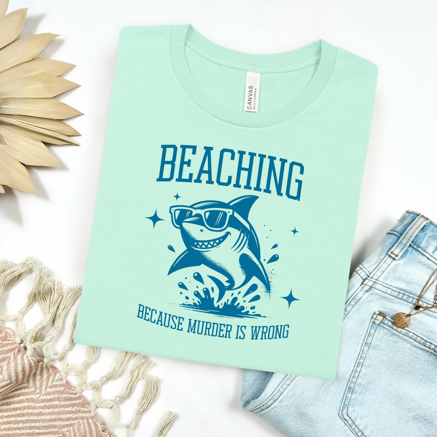 Bella Canvas 3001 Beaching Because Murder Is Wrong T Shirt with Funny Shark with Sunglasses splashing in water in one color graphic. Picture on a flay lay mockup with pair of blue jeans and blanket and a wicker sun shaped accessory.