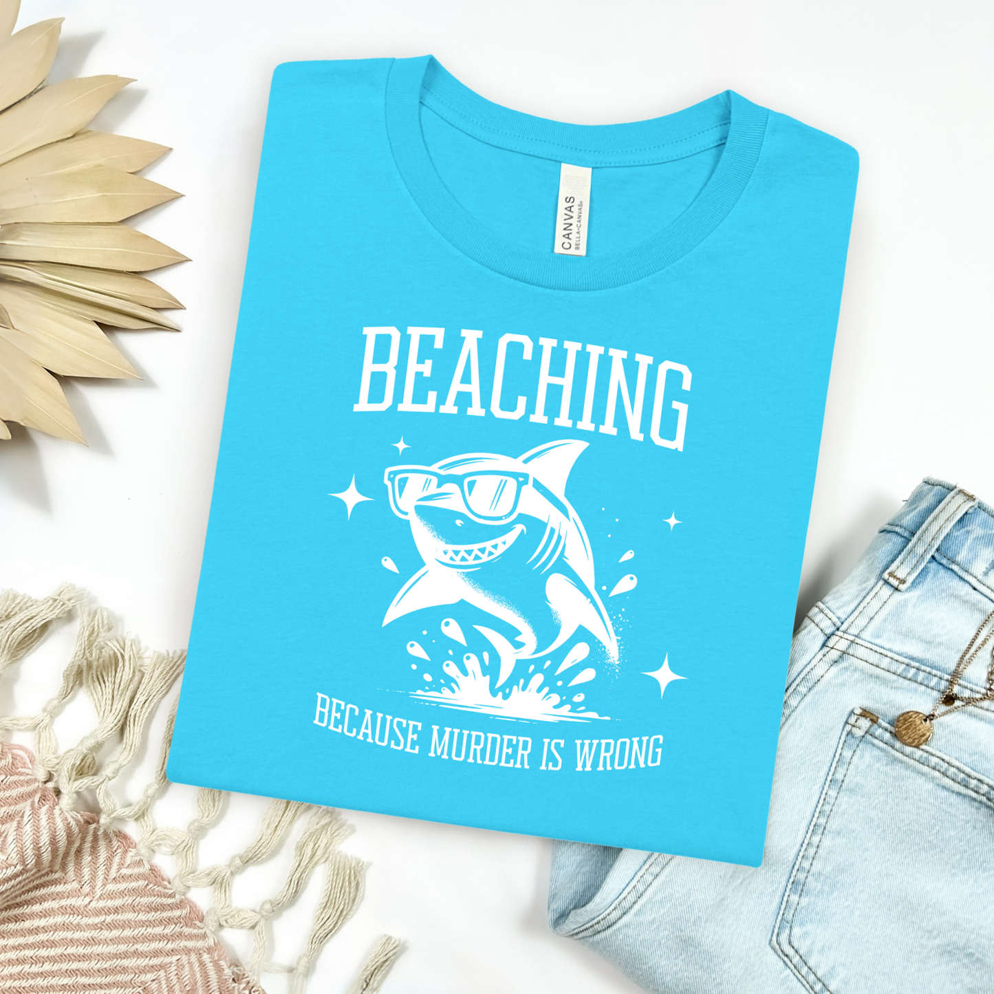 Bella Canvas 3001 Beaching Because Murder Is Wrong T Shirt with Funny Shark with Sunglasses splashing in water in one color graphic. Picture on a flay lay mockup with pair of blue jeans and blanket and a wicker sun shaped accessory.