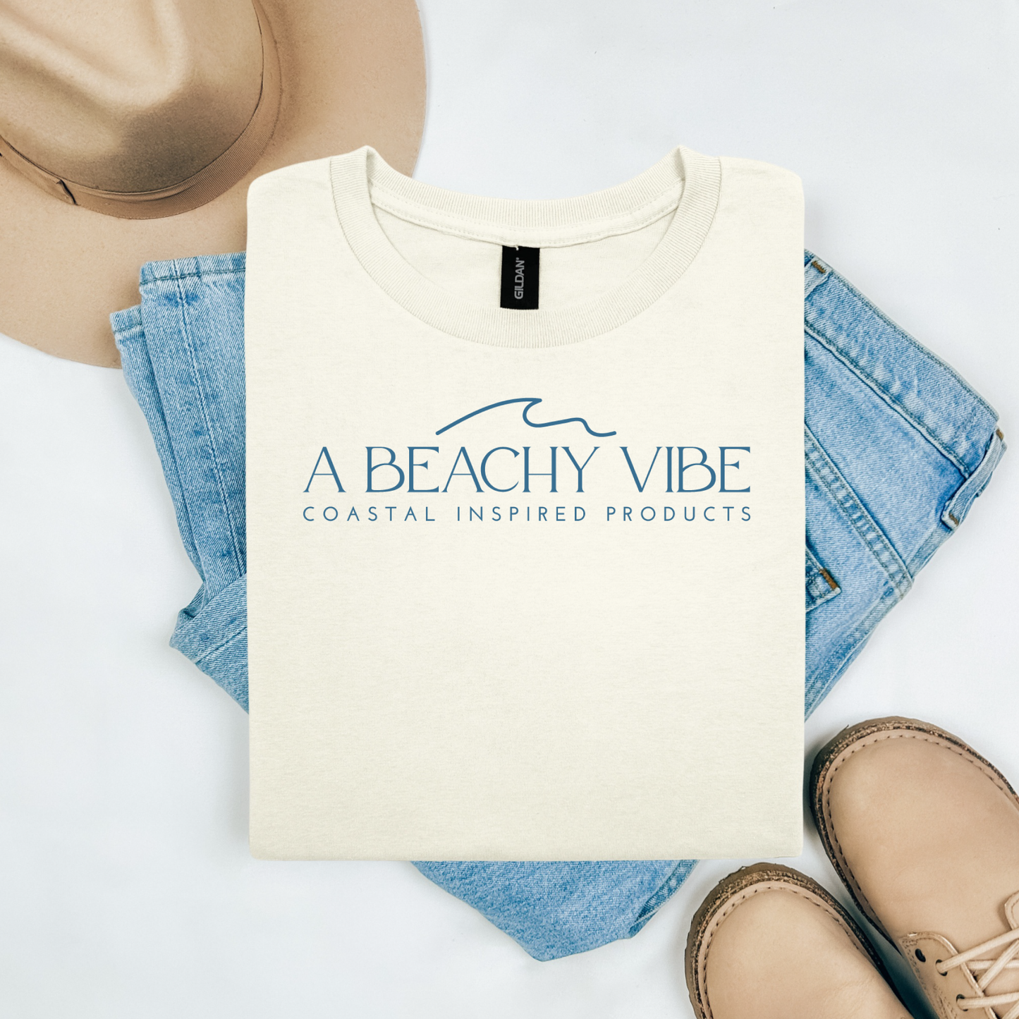 Gildan 5000 "A Beachy Vibe" t shirt with a simple wave graphic in one color. Picture on a flay lay mockup with pair of blue jeans, brown shoes and a hat accessory.