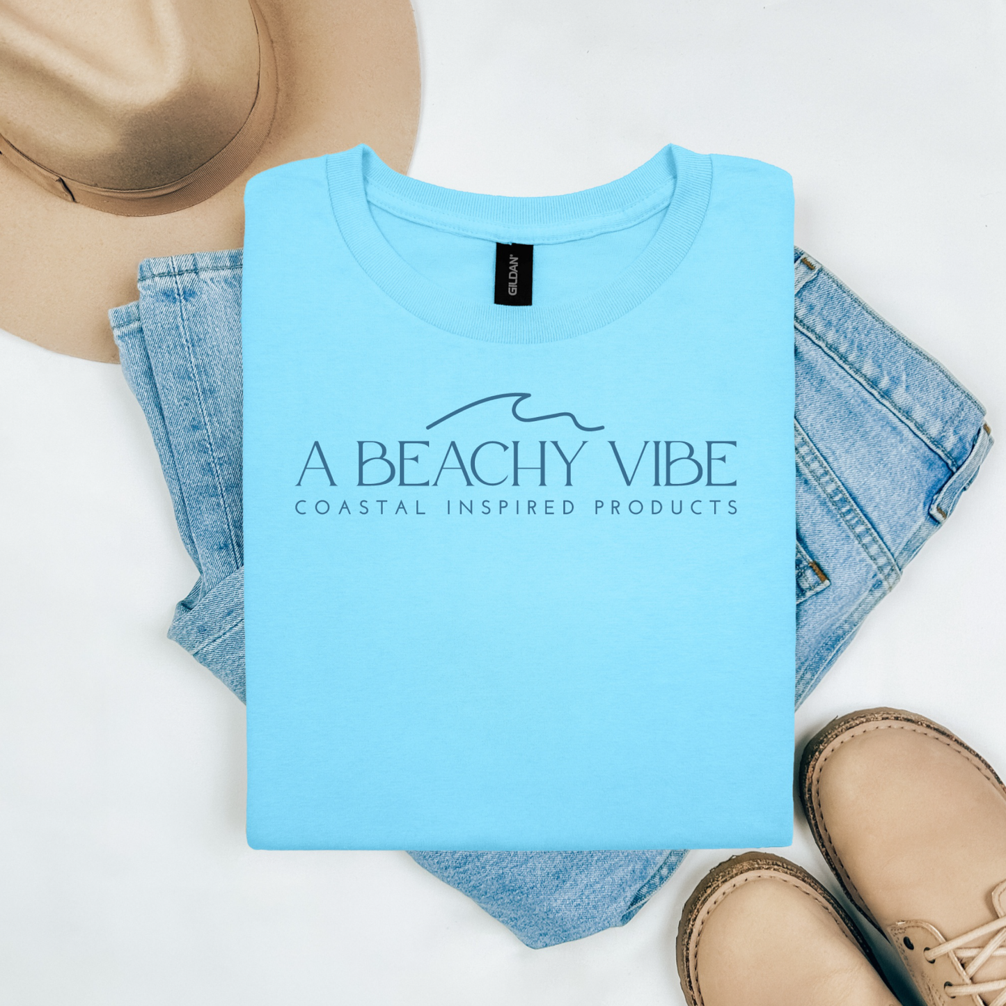 Gildan 5000 "A Beachy Vibe" t shirt with a simple wave graphic in one color. Picture on a flay lay mockup with pair of blue jeans, brown shoes and a hat accessory.