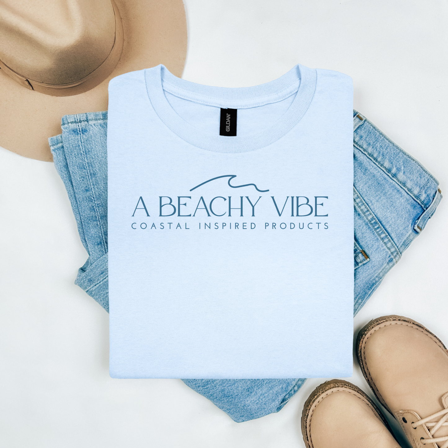 Gildan 5000 "A Beachy Vibe" t shirt with a simple wave graphic in one color. Picture on a flay lay mockup with pair of blue jeans, brown shoes and a hat accessory.