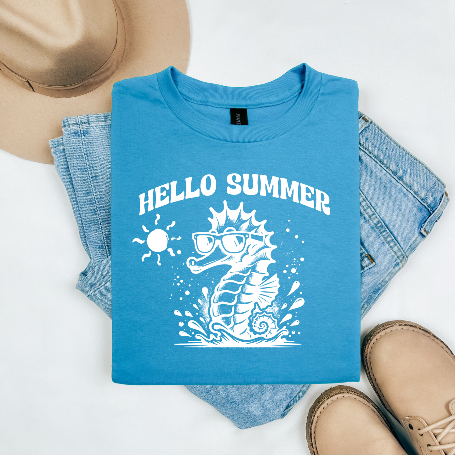 Gildan 5000 "Hello Summer" t shirt with a cute seahorse with sunglasses in one color graphics. Picture on a flay lay mockup with pair of blue jeans, brown shoes and a hat accessory.