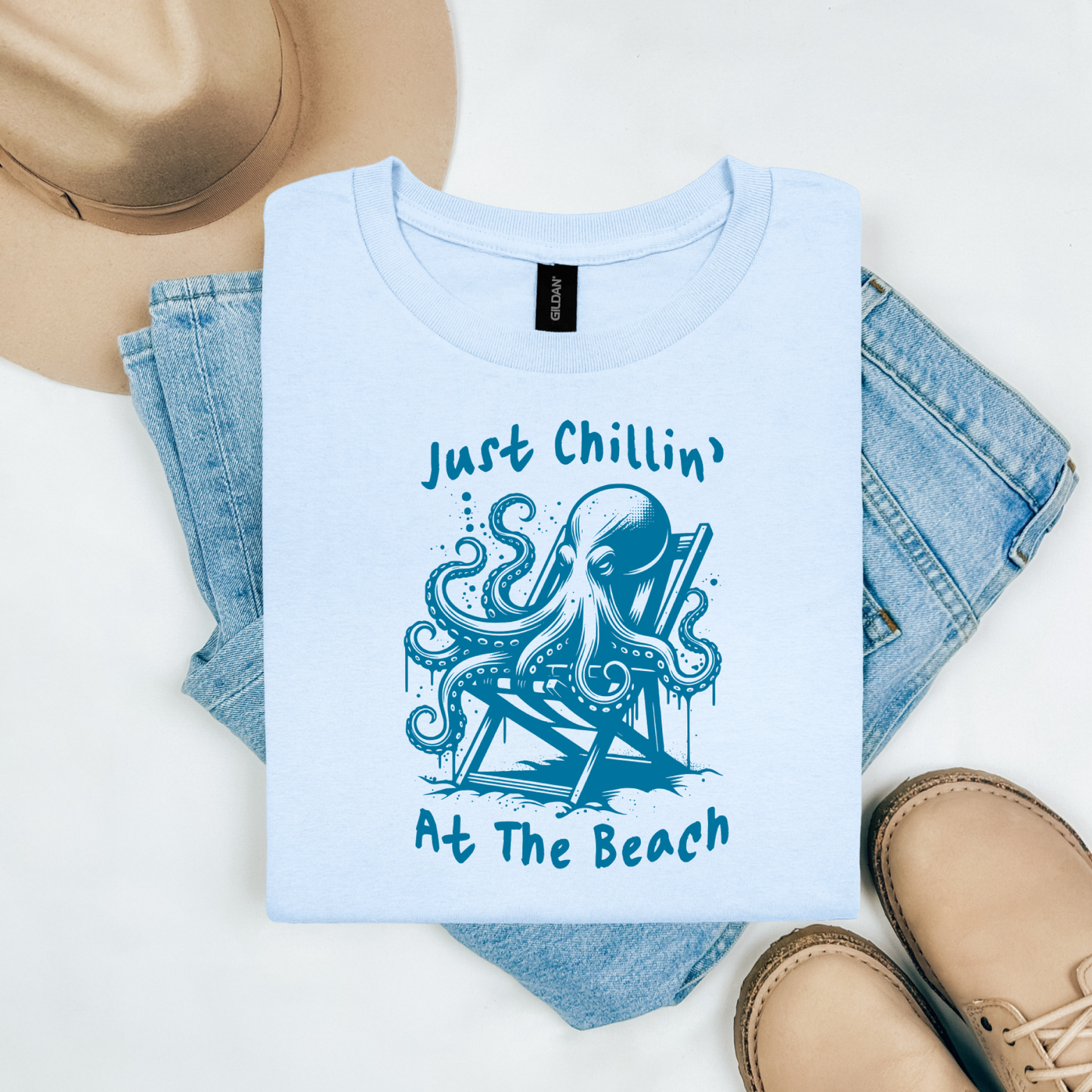 Gildan 5000 "Just Chillin' At The Beach" t shirt with an octopus setting in a beach chair in one color graphics. Picture on a flay lay mockup with pair of blue jeans, brown shoes and a hat accessory.