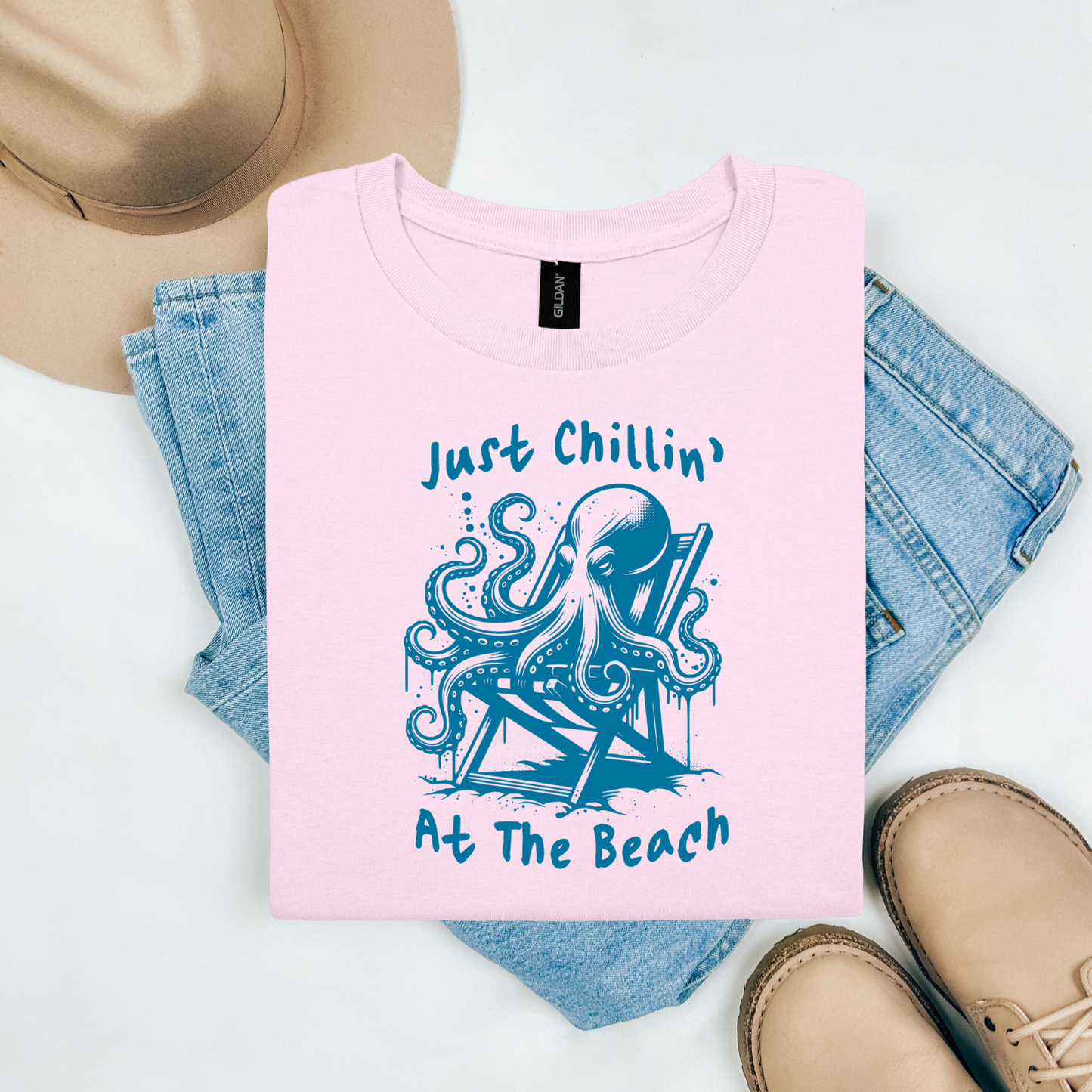 Gildan 5000 "Just Chillin' At The Beach" t shirt with an octopus setting in a beach chair in one color graphics. Picture on a flay lay mockup with pair of blue jeans, brown shoes and a hat accessory.