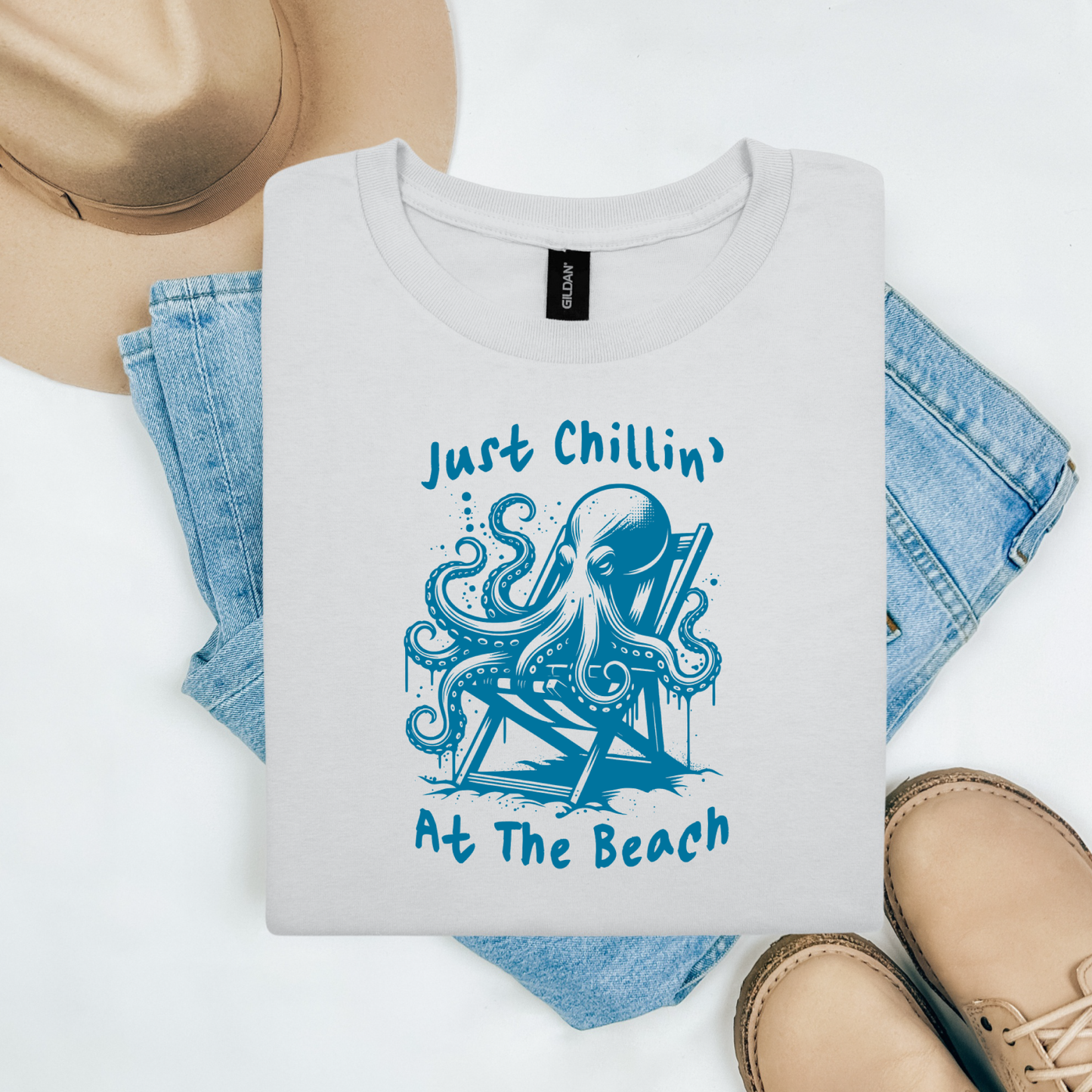 Gildan 5000 "Just Chillin' At The Beach" t shirt with an octopus setting in a beach chair in one color graphics. Picture on a flay lay mockup with pair of blue jeans, brown shoes and a hat accessory.