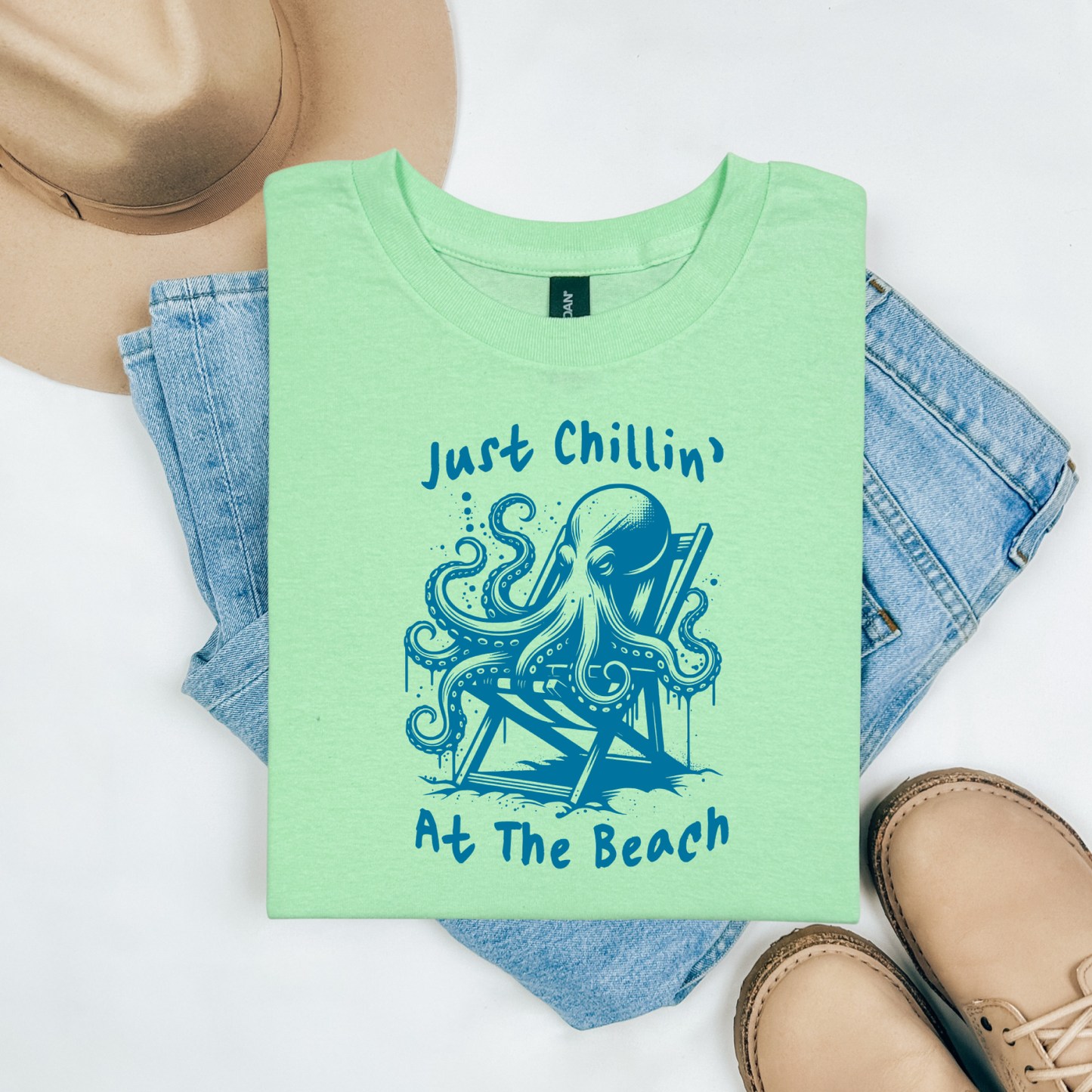 Gildan 5000 "Just Chillin' At The Beach" t shirt with an octopus setting in a beach chair in one color graphics. Picture on a flay lay mockup with pair of blue jeans, brown shoes and a hat accessory.
