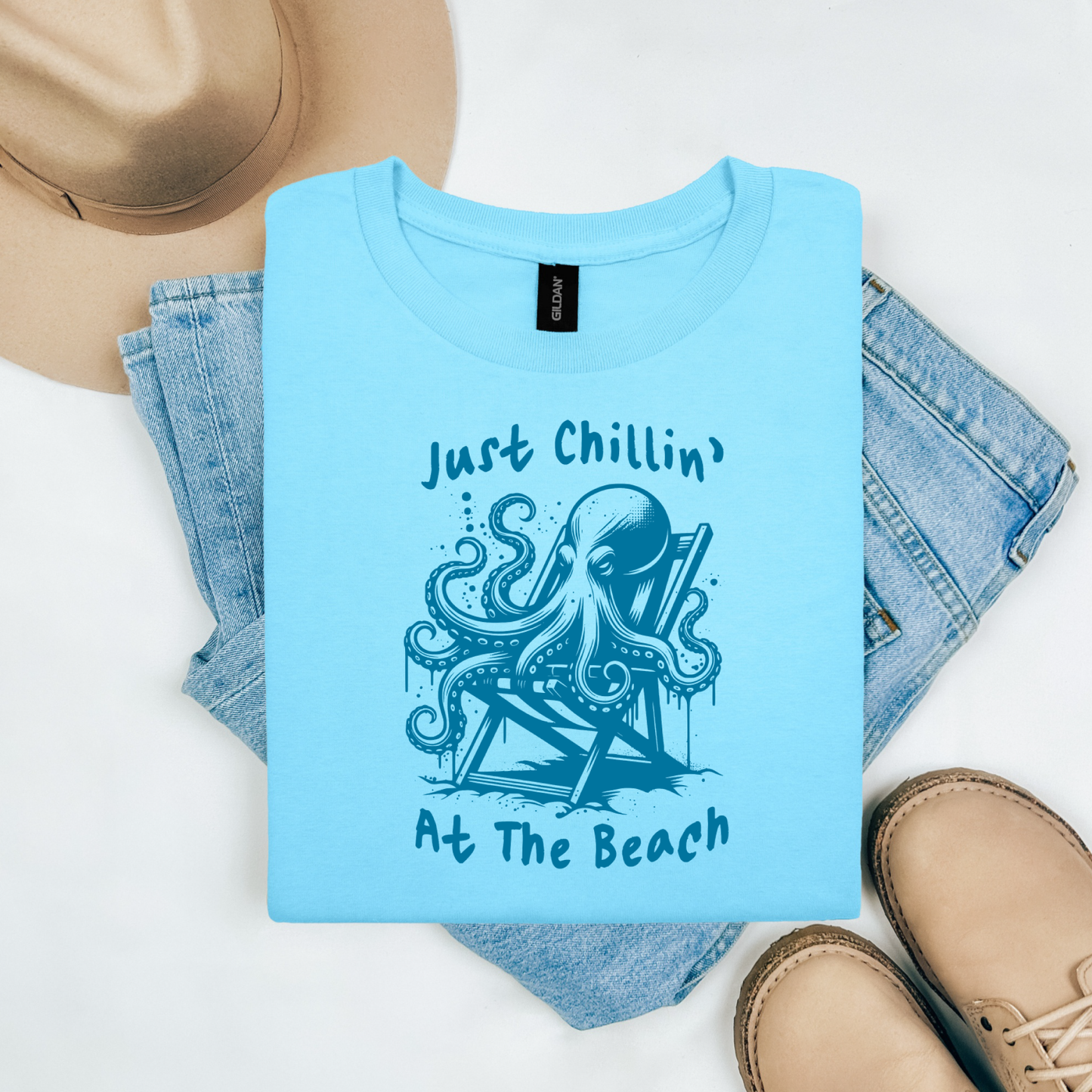 Gildan 5000 "Just Chillin' At The Beach" t shirt with an octopus setting in a beach chair in one color graphics. Picture on a flay lay mockup with pair of blue jeans, brown shoes and a hat accessory.
