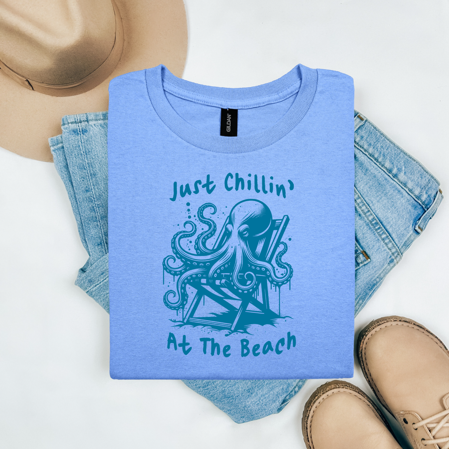 Gildan 5000 "Just Chillin' At The Beach" t shirt with an octopus setting in a beach chair in one color graphics. Picture on a flay lay mockup with pair of blue jeans, brown shoes and a hat accessory.