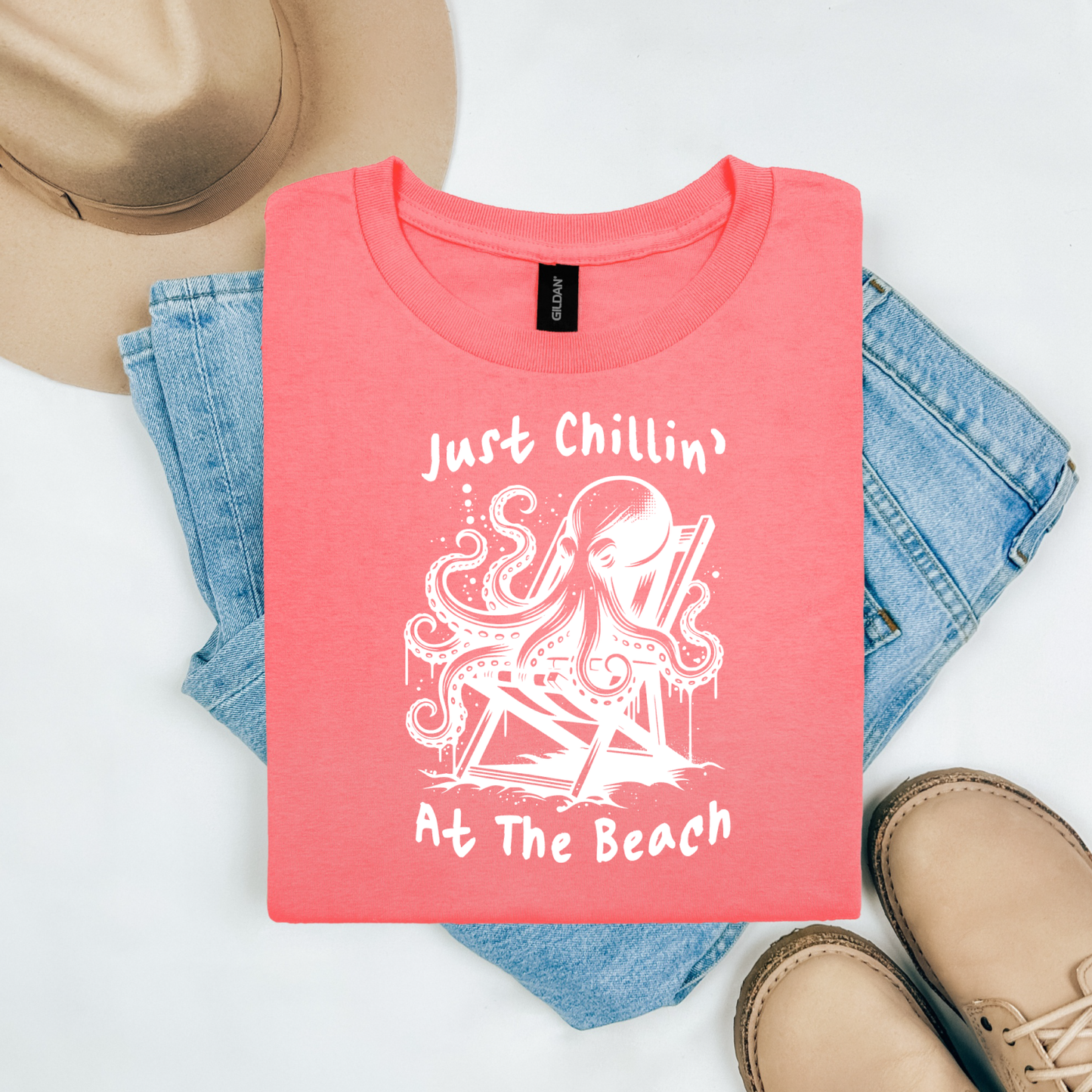 Gildan 5000 "Just Chillin' At The Beach" t shirt with an octopus setting in a beach chair in one color graphics. Picture on a flay lay mockup with pair of blue jeans, brown shoes and a hat accessory.