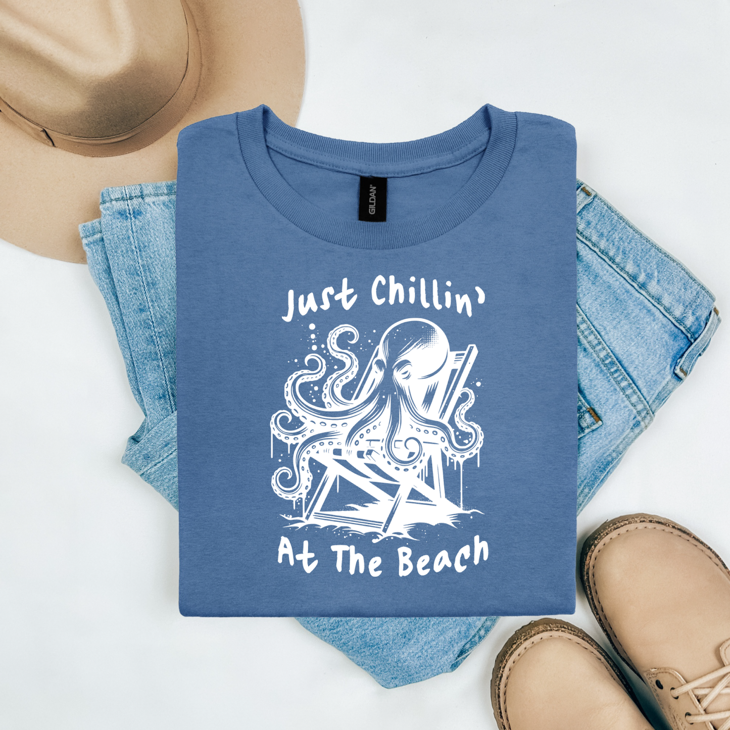 Gildan 5000 "Just Chillin' At The Beach" t shirt with an octopus setting in a beach chair in one color graphics. Picture on a flay lay mockup with pair of blue jeans, brown shoes and a hat accessory.