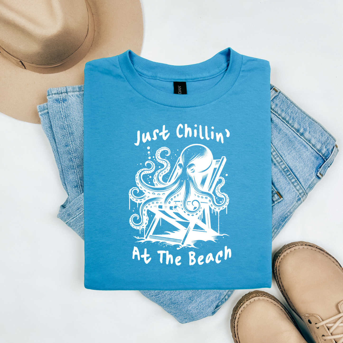 Gildan 5000 "Just Chillin' At The Beach" t shirt with an octopus setting in a beach chair in one color graphics. Picture on a flay lay mockup with pair of blue jeans, brown shoes and a hat accessory.