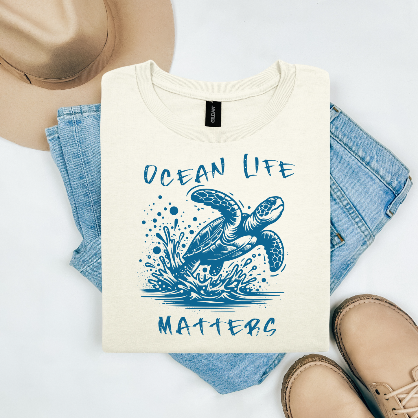Gildan 5000 "Save The Ocean and The Sea Life In It" t shirt with a sea turtle splashing in water in a one color graphic. Picture on a flay lay mockup with pair of blue jeans, brown shoes and a hat accessory.