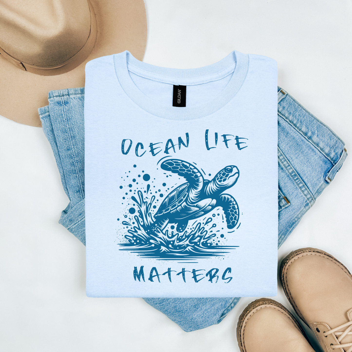 Gildan 5000 "Save The Ocean and The Sea Life In It" t shirt with a sea turtle splashing in water in a one color graphic. Picture on a flay lay mockup with pair of blue jeans, brown shoes and a hat accessory.