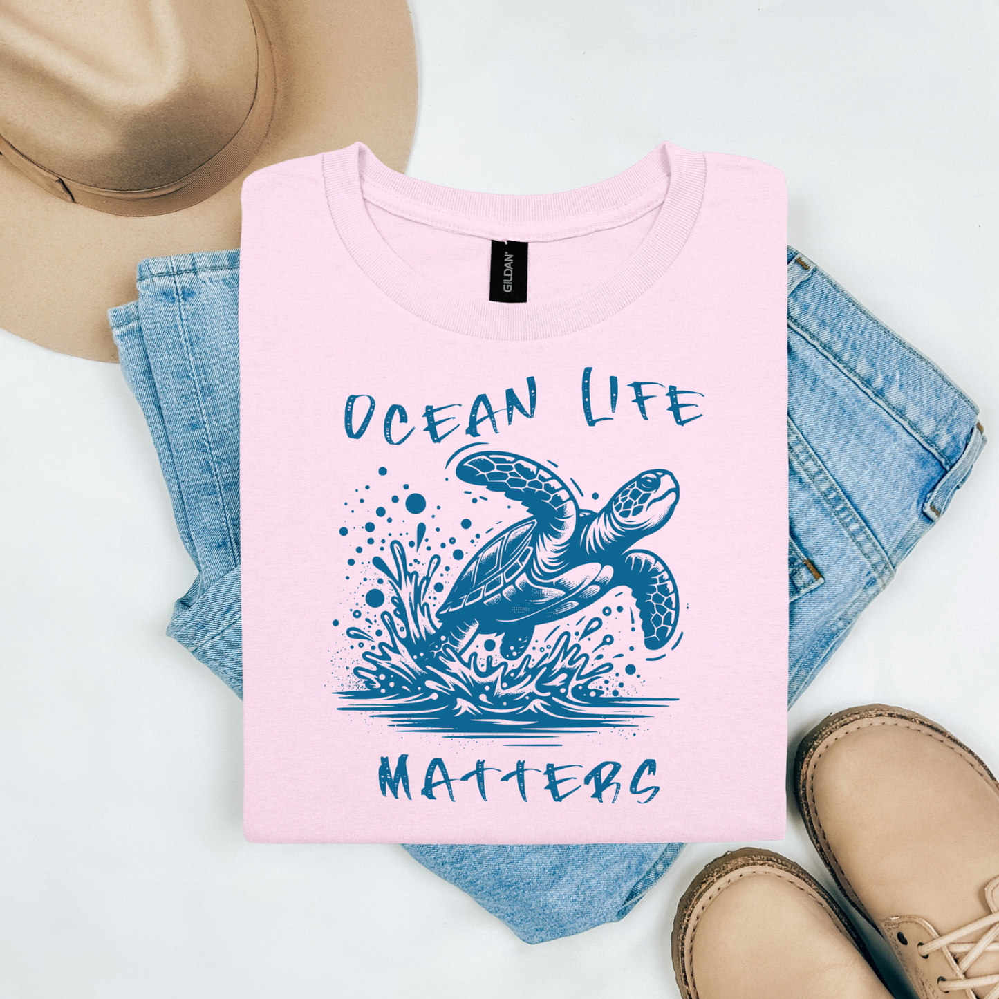 Gildan 5000 "Save The Ocean and The Sea Life In It" t shirt with a sea turtle splashing in water in a one color graphic. Picture on a flay lay mockup with pair of blue jeans, brown shoes and a hat accessory.
