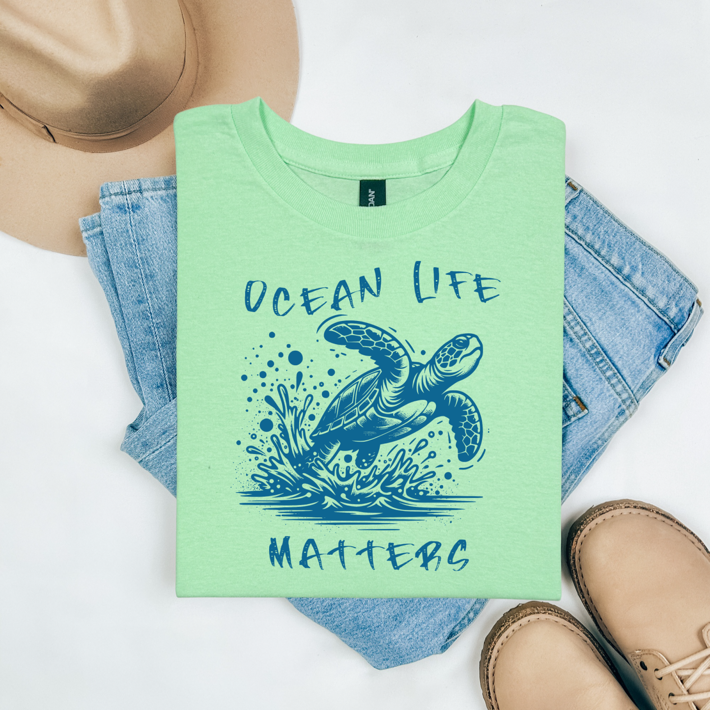 Gildan 5000 "Save The Ocean and The Sea Life In It" t shirt with a sea turtle splashing in water in a one color graphic. Picture on a flay lay mockup with pair of blue jeans, brown shoes and a hat accessory.