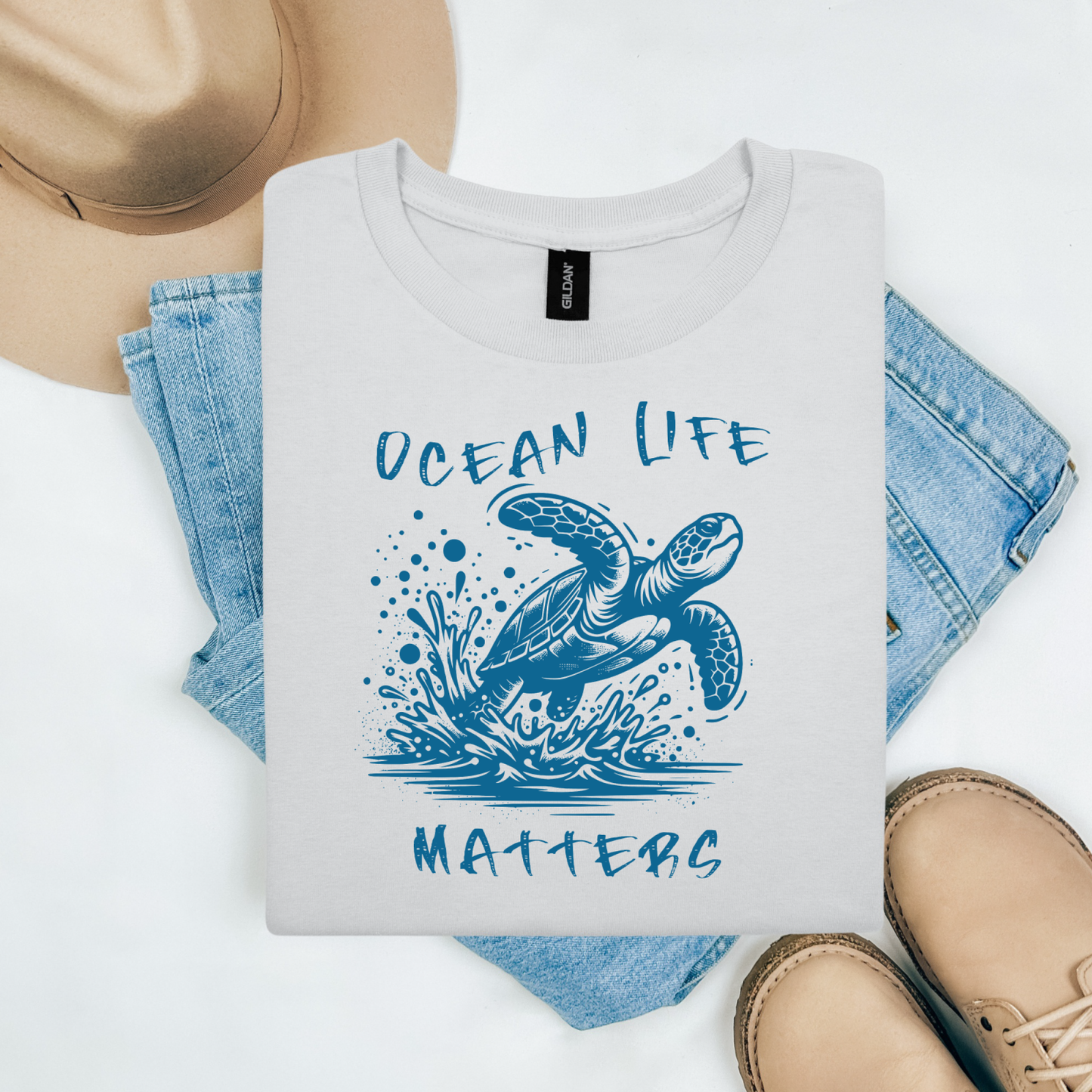 Gildan 5000 "Save The Ocean and The Sea Life In It" t shirt with a sea turtle splashing in water in a one color graphic. Picture on a flay lay mockup with pair of blue jeans, brown shoes and a hat accessory.