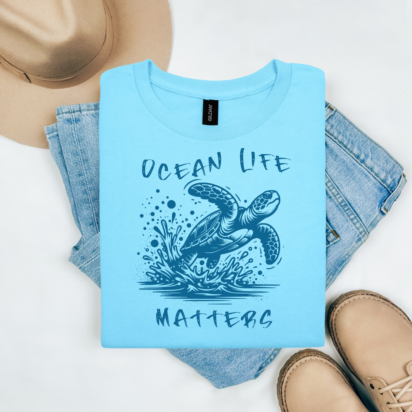 Gildan 5000 "Save The Ocean and The Sea Life In It" t shirt with a sea turtle splashing in water in a one color graphic. Picture on a flay lay mockup with pair of blue jeans, brown shoes and a hat accessory.
