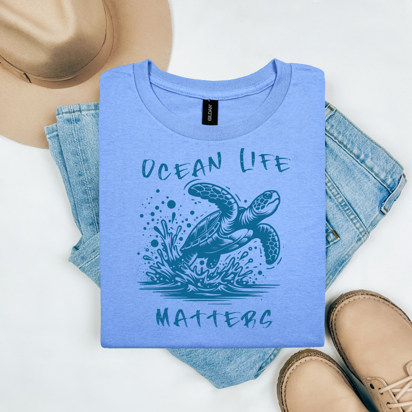 Gildan 5000 "Save The Ocean and The Sea Life In It" t shirt with a sea turtle splashing in water in a one color graphic. Picture on a flay lay mockup with pair of blue jeans, brown shoes and a hat accessory.