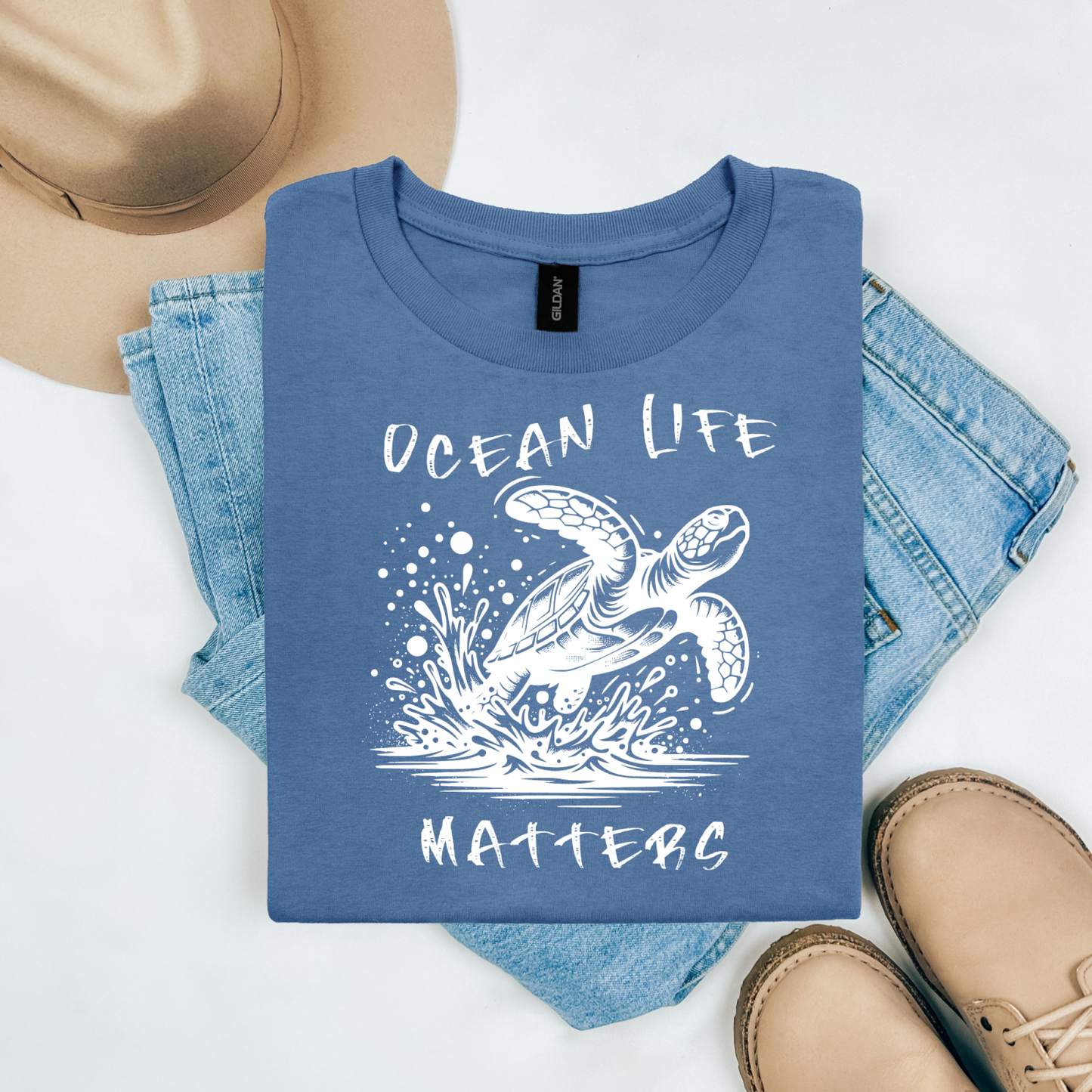 Gildan 5000 "Save The Ocean and The Sea Life In It" t shirt with a sea turtle splashing in water in a one color graphic. Picture on a flay lay mockup with pair of blue jeans, brown shoes and a hat accessory.